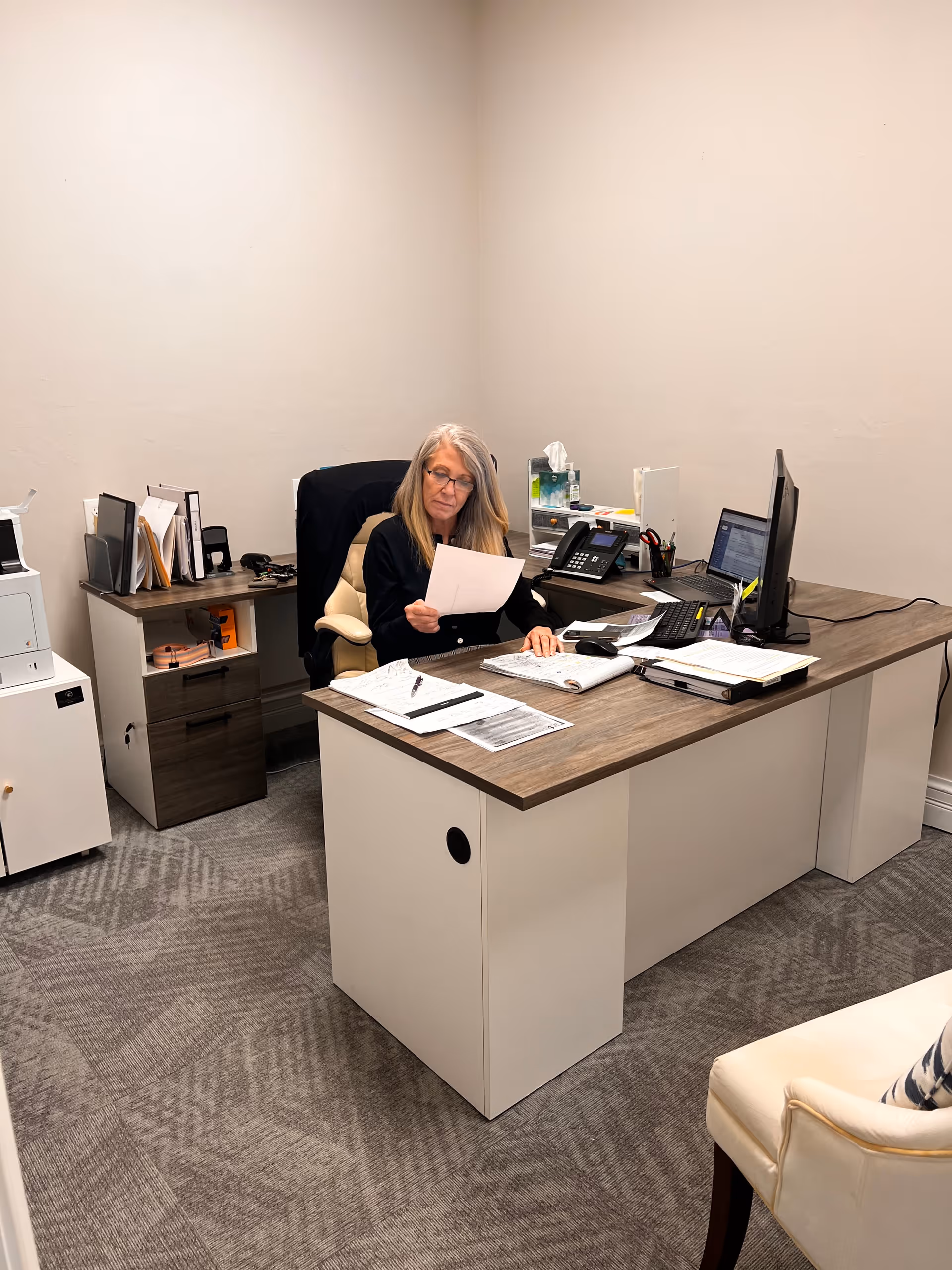 A woman sits at a wood desk in a small office, reviewing documents.