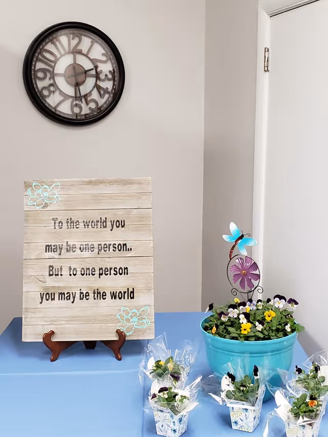 Interior wall scene with a clock, inspirational wooden sign, and potted flowers on a blue table.