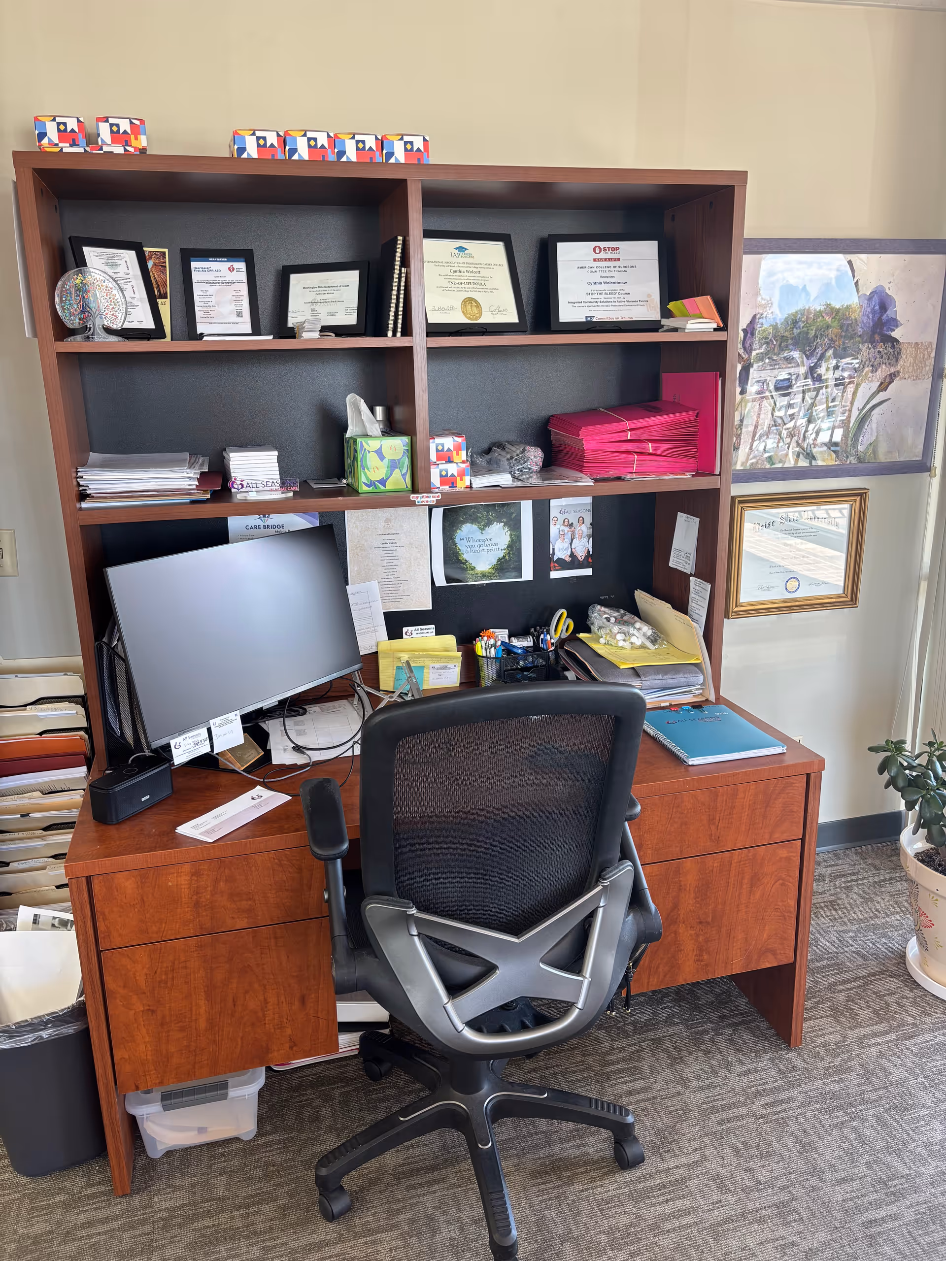 A wooden desk and shelving unit with certificates, a computer, and office supplies in a facility.