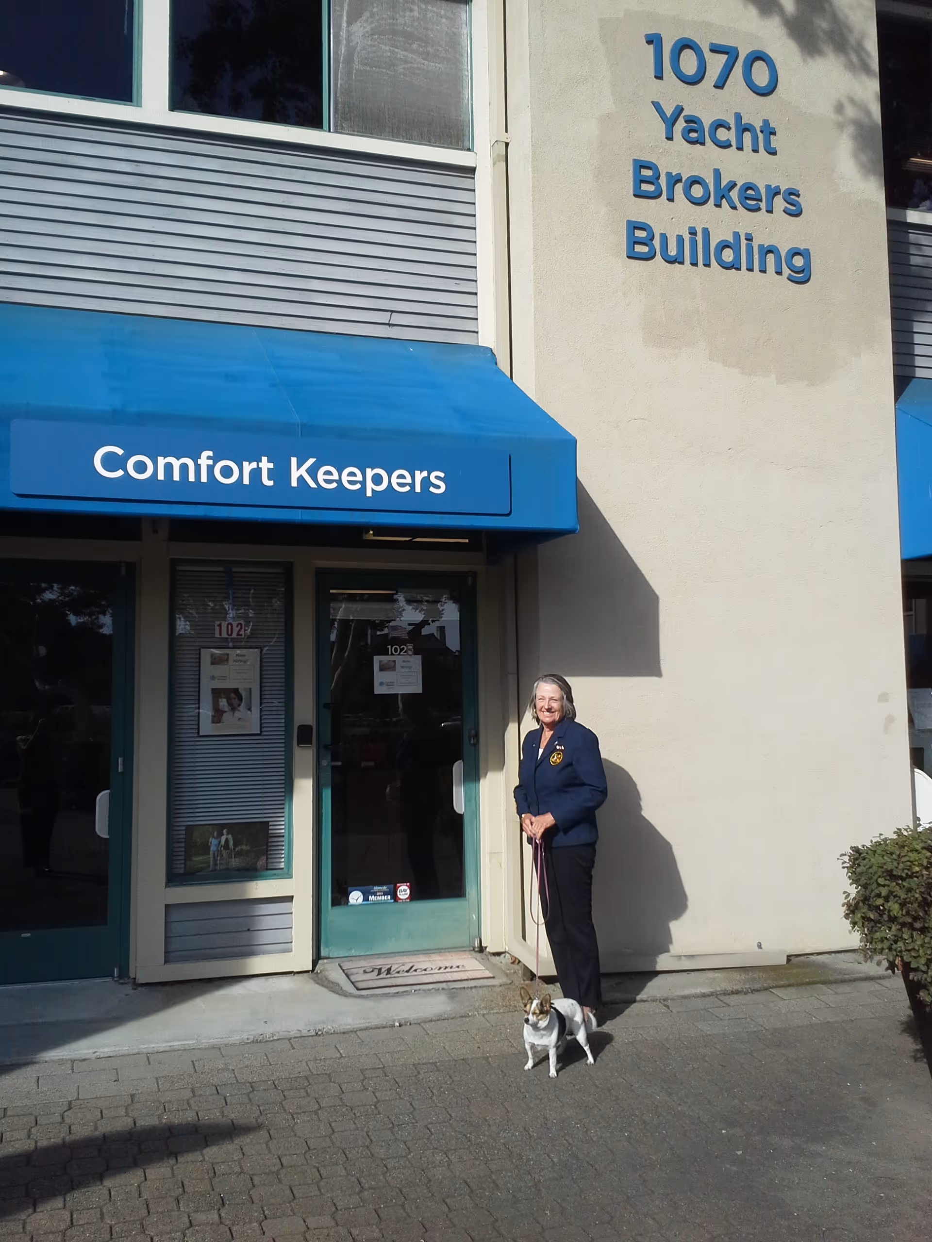 Exterior of Comfort Keepers storefront with blue canopy, a person and dog outside.