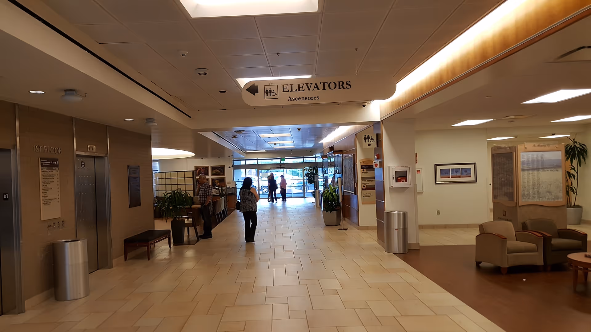 Hospital lobby interior with elevators, seating, plants, and visitors walking toward the entrance.