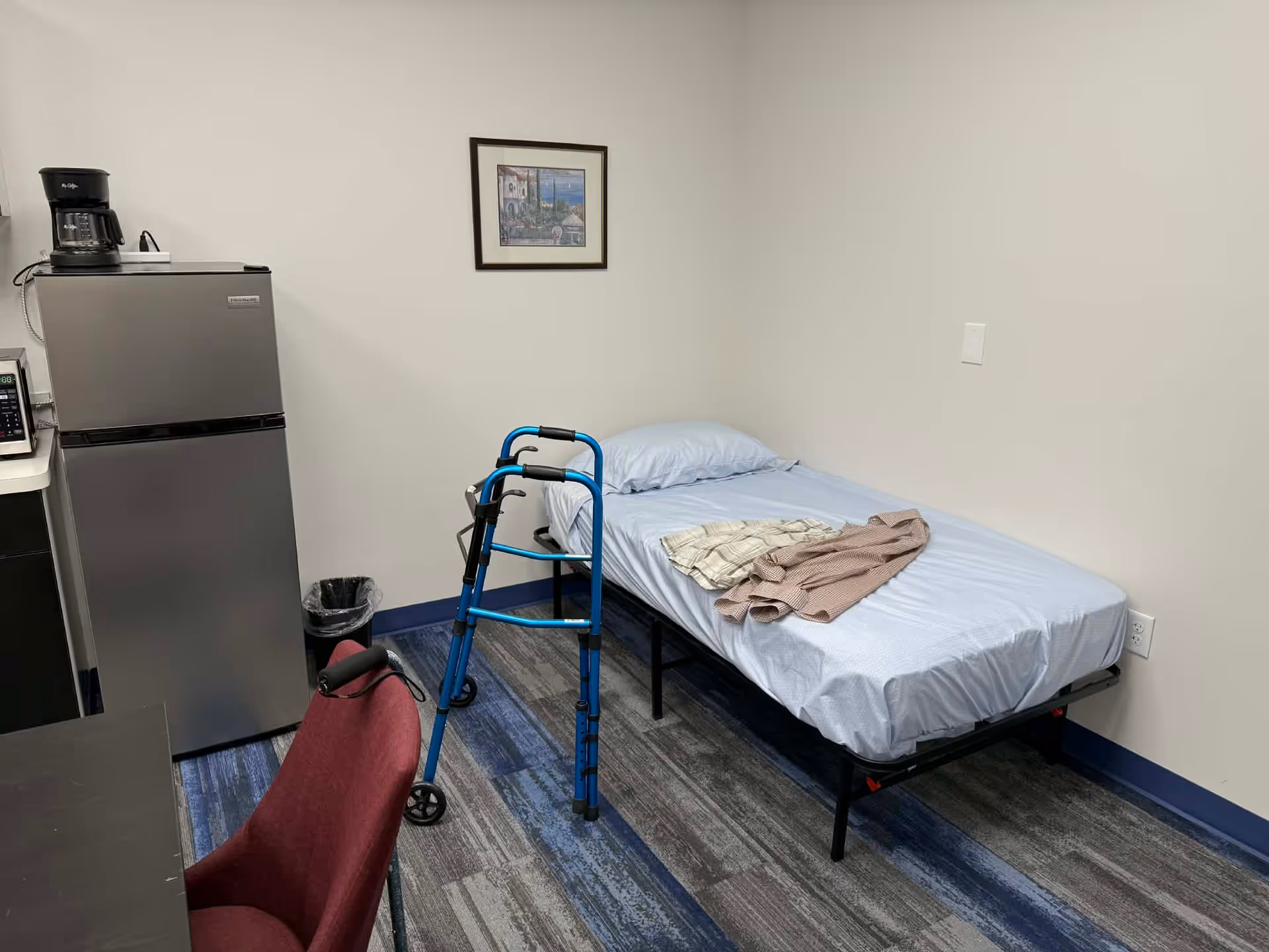 Small senior living bedroom with single bed, blue sheets, walker, chair, and small fridge.
