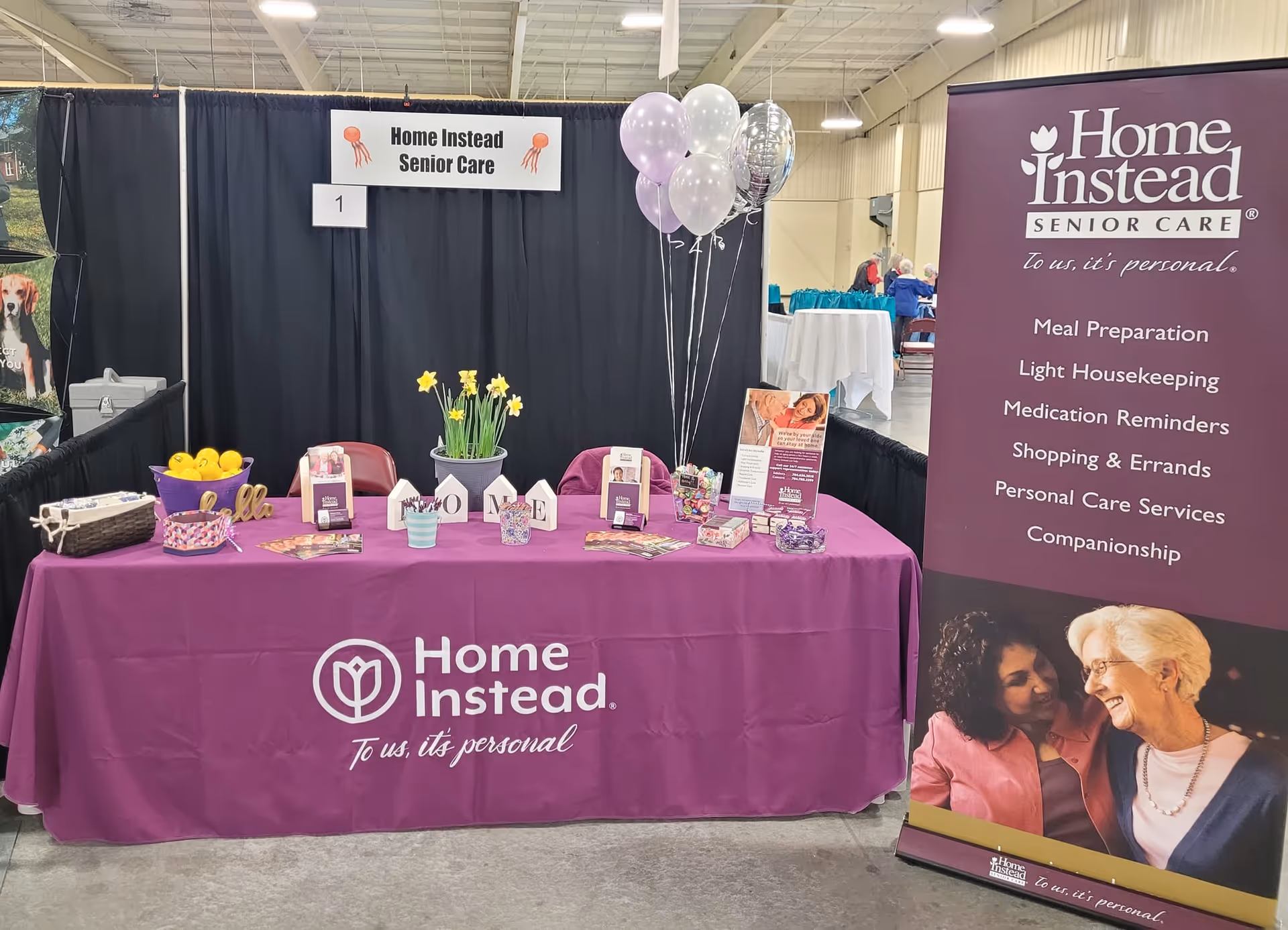 Indoor Home Instead booth with purple tablecloth, balloons, and promotional materials at expo.