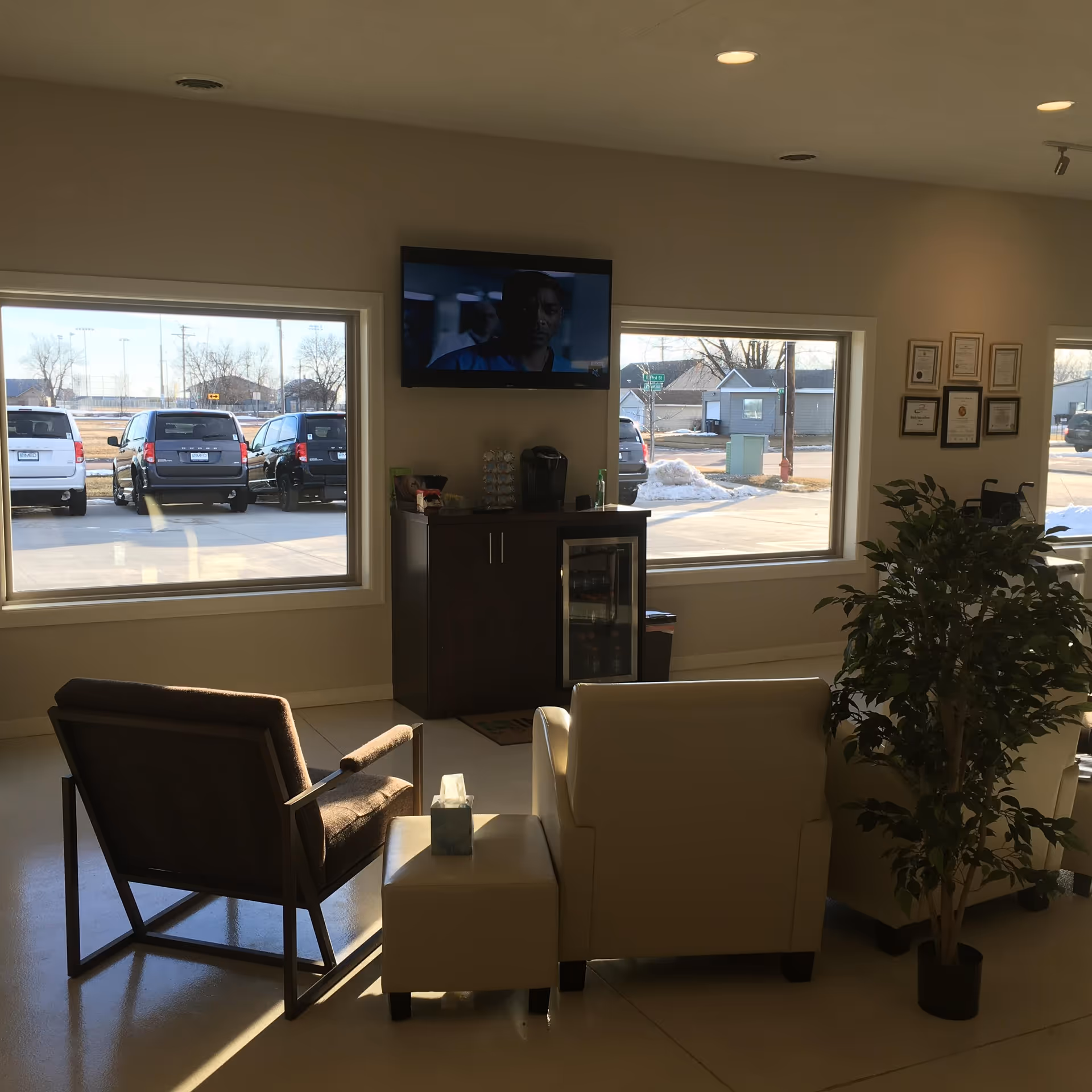 A modern senior living lounge with seating, a TV, a plant, and large windows.