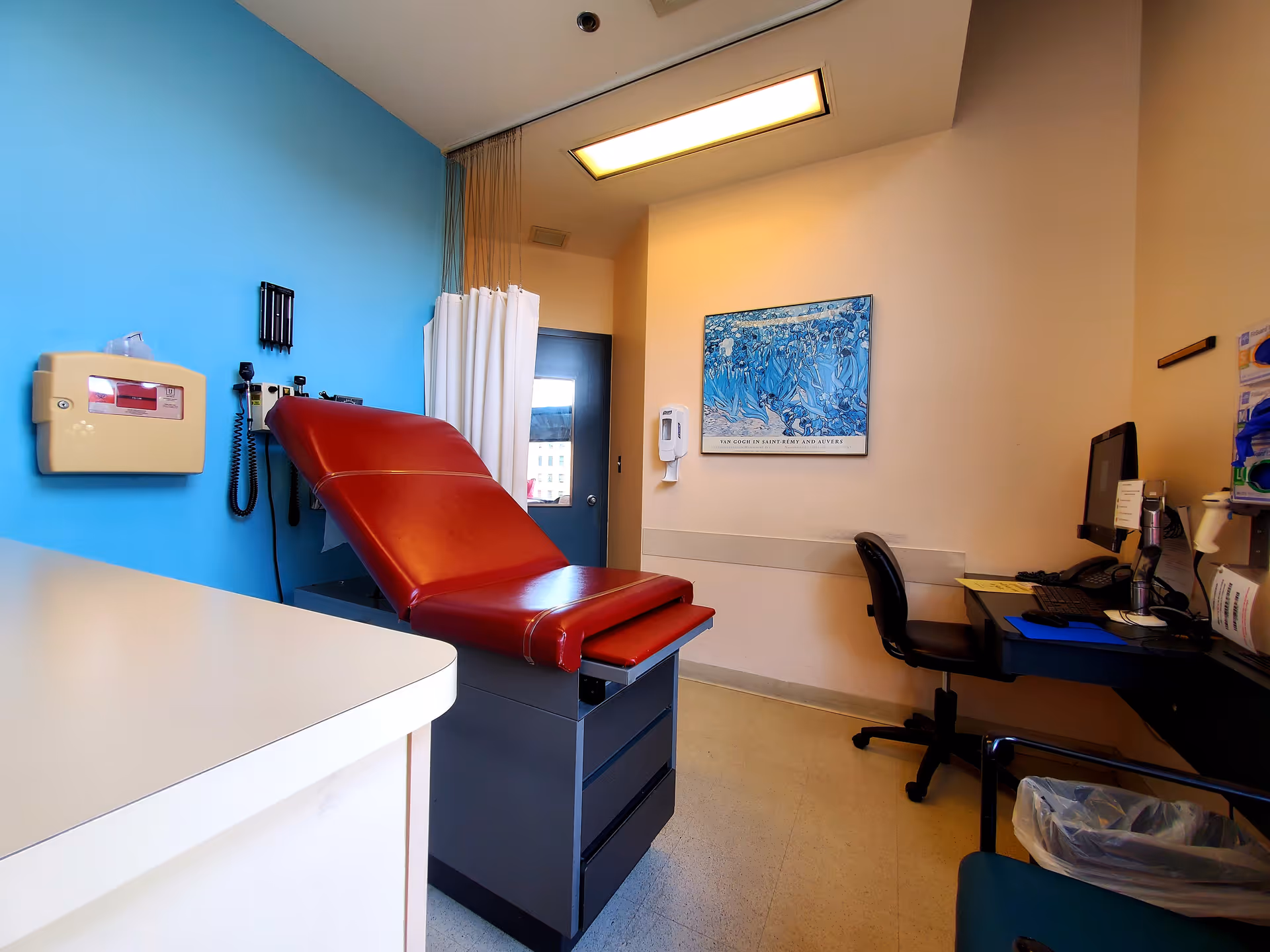 A red medical examination chair sits in a bright clinic exam room.