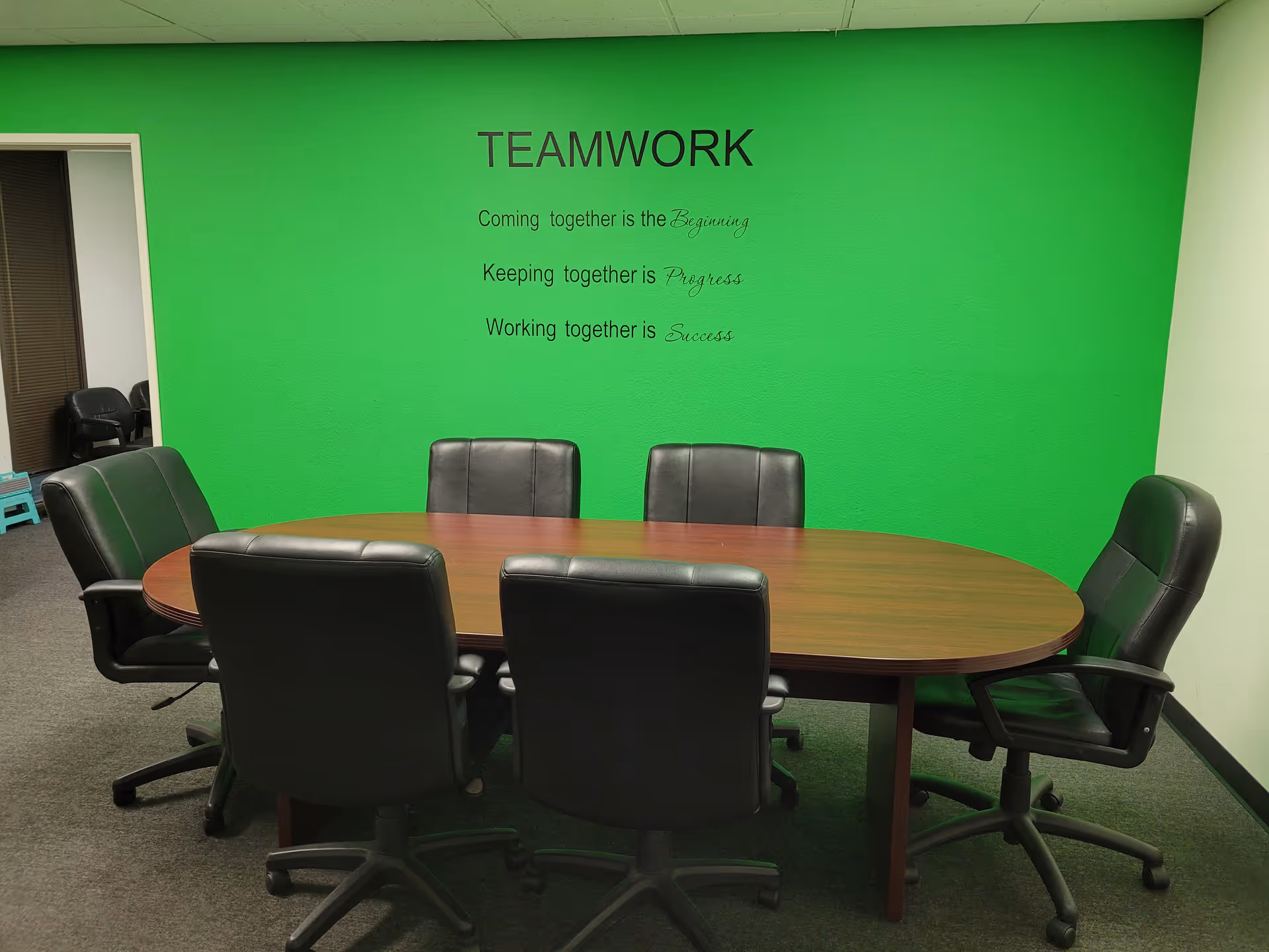 Bright green-walled conference room with oval table and six black leather chairs.