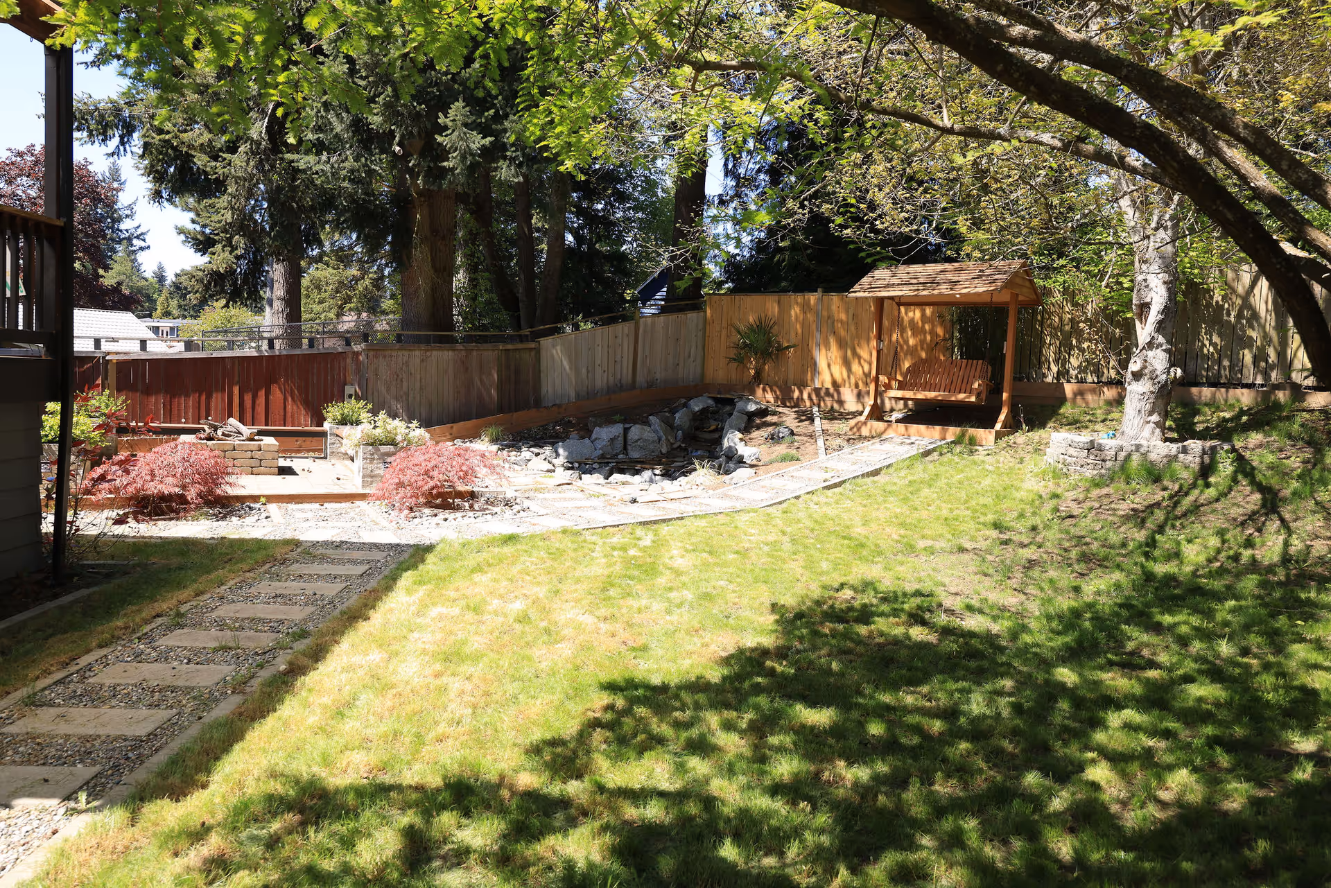 Backyard garden with wooden gazebo swing, stone path, trees, and sunlit lawn.