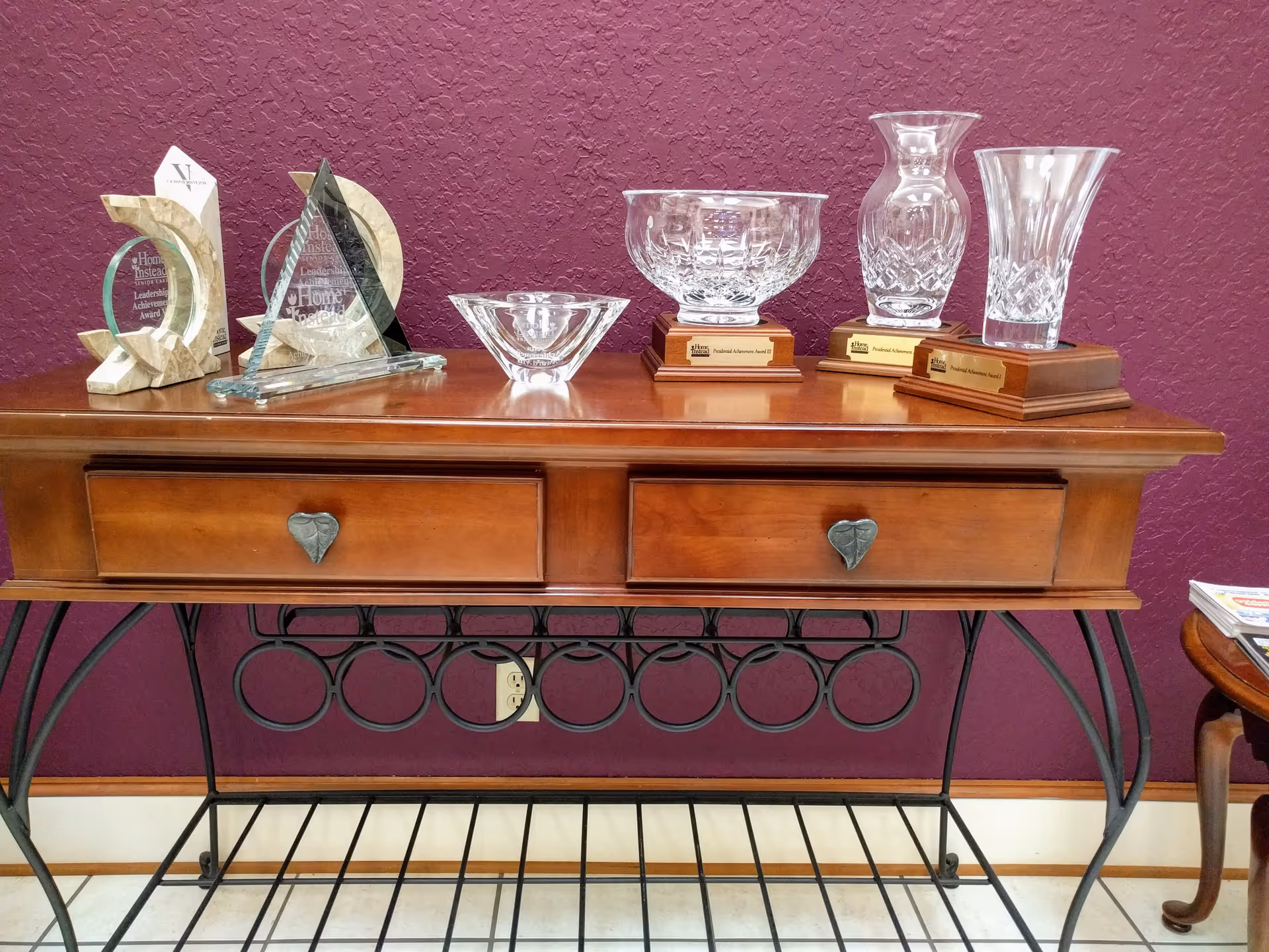 Wooden console table against a purple wall displays glass trophies and plaques.