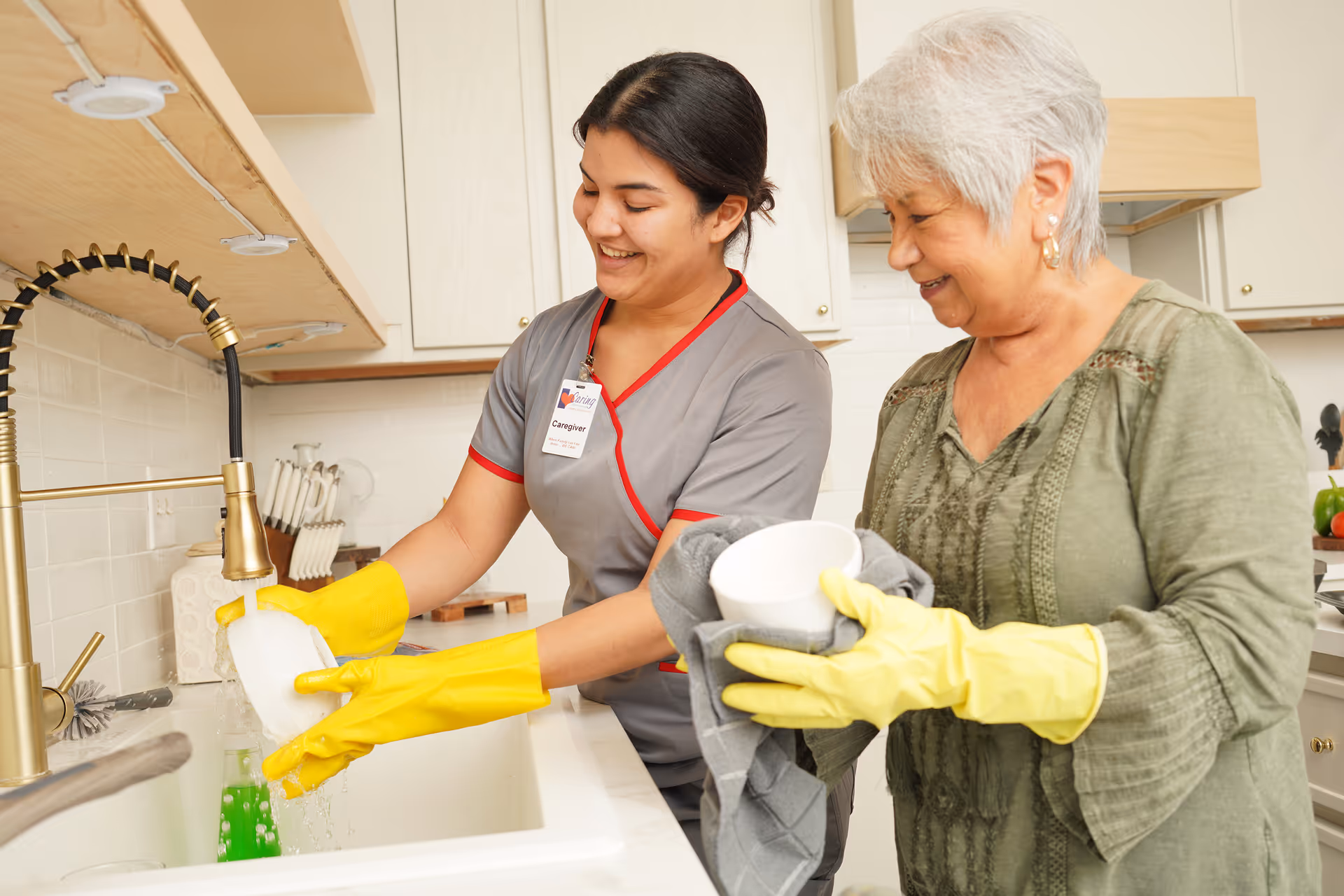 Caregiver and elderly woman washing dishes at a kitchen sink, both wearing yellow gloves.