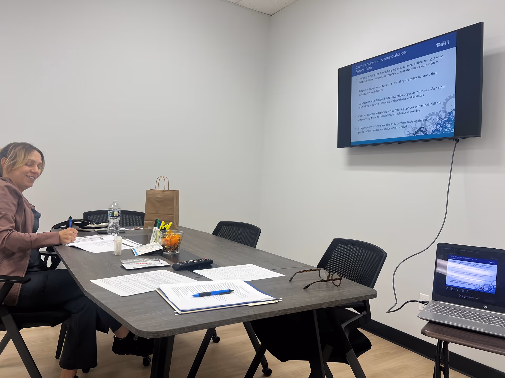 A woman sits at a conference table with papers, a laptop, and a wall-mounted screen.
