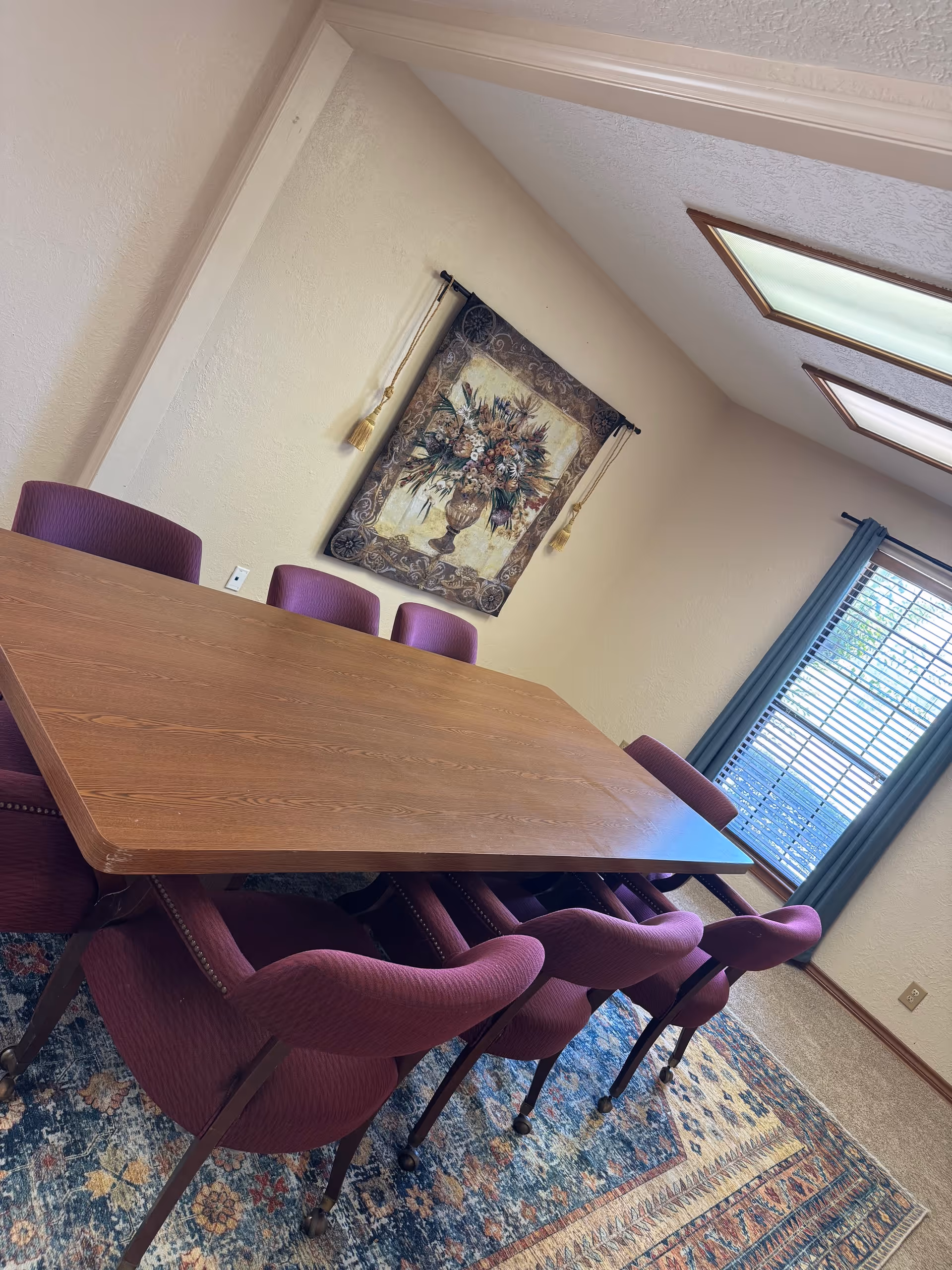 A wooden dining table with purple upholstered chairs in a bright, carpeted room.