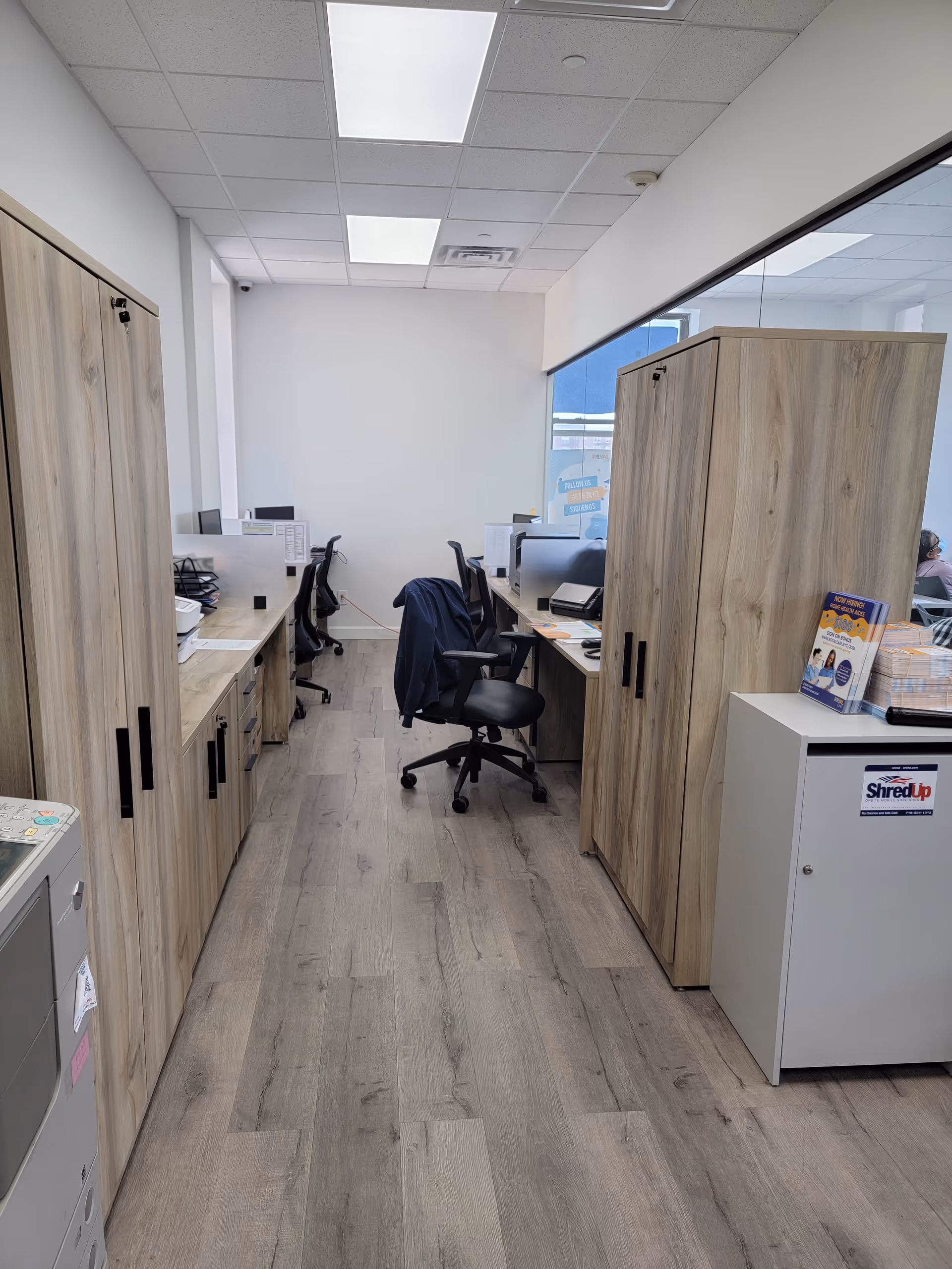 Narrow office corridor with desks, chairs, and tall wooden cabinets in an administrative area.