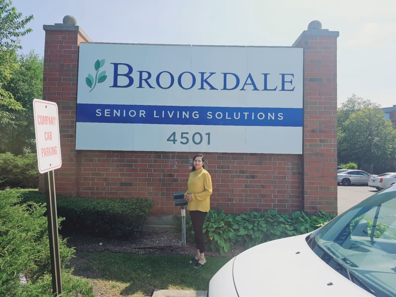 A person stands beside a Brookdale Senior Living sign at an exterior facility entrance.