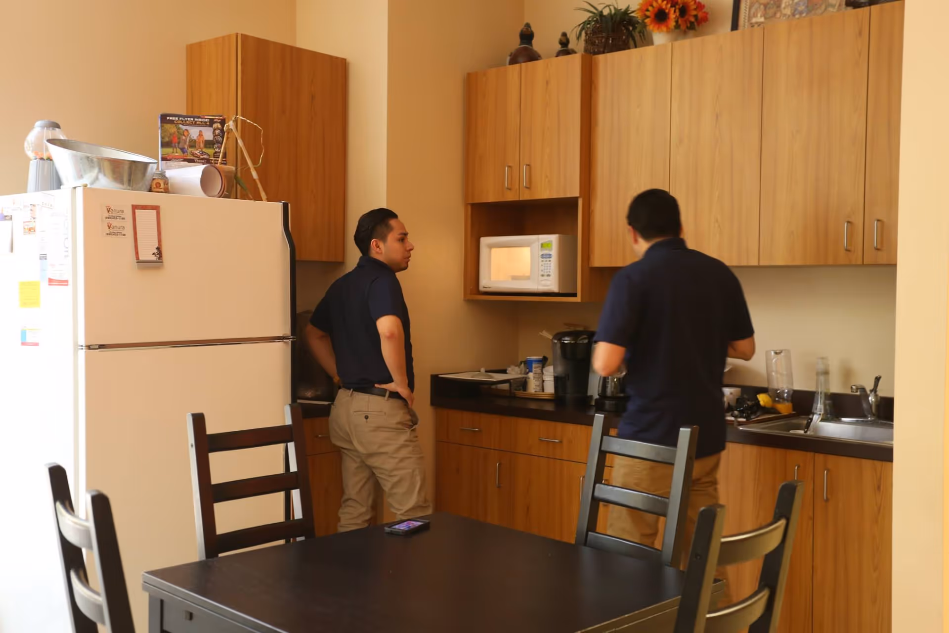 Two staff members stand in a kitchenette with a fridge, microwave, and wooden cabinets.