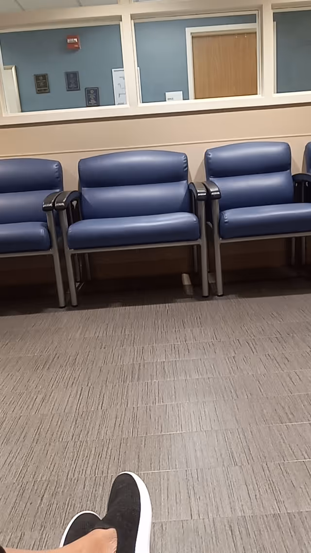 Blue waiting area chairs line a hallway inside a facility, with a person's feet visible.