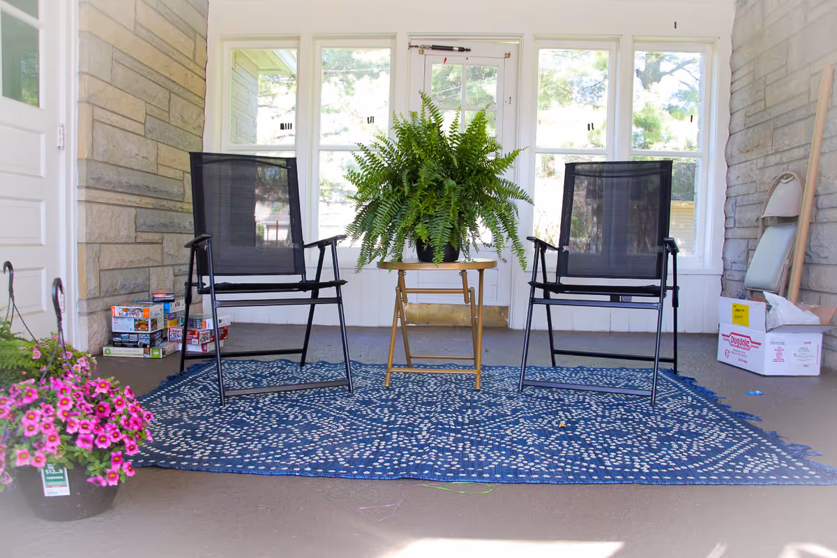 Two black patio chairs flank a fern on a small wooden table on a covered porch.
