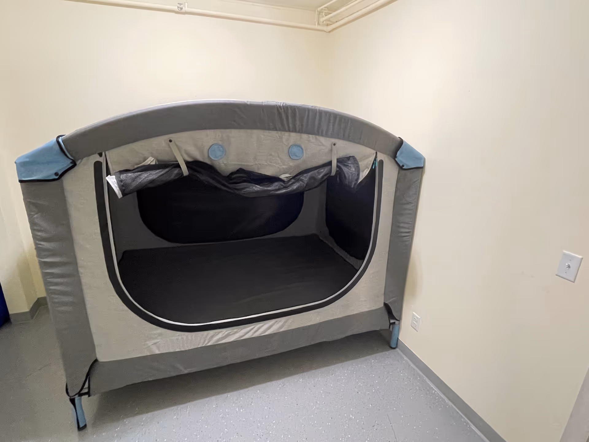 Gray portable bed enclosure with open front in a small, plain room.