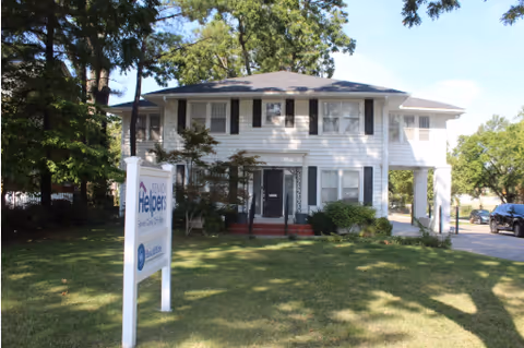 Two-story white building with Senior Helpers sign, front lawn, and trees.