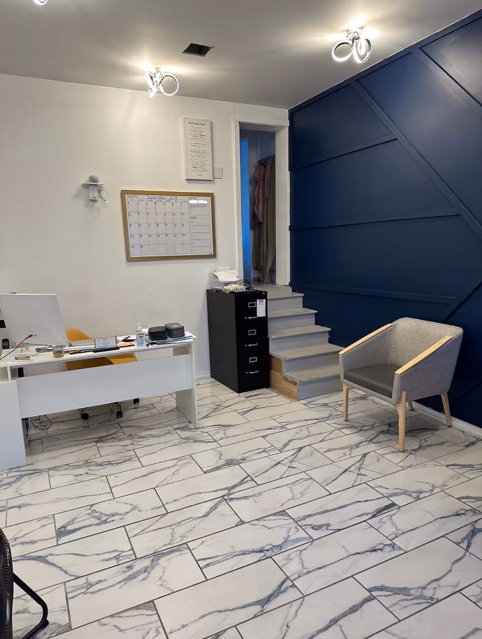 Office reception area with white desk, calendar, filing cabinet, stairway, blue wall paneling, marble floor.