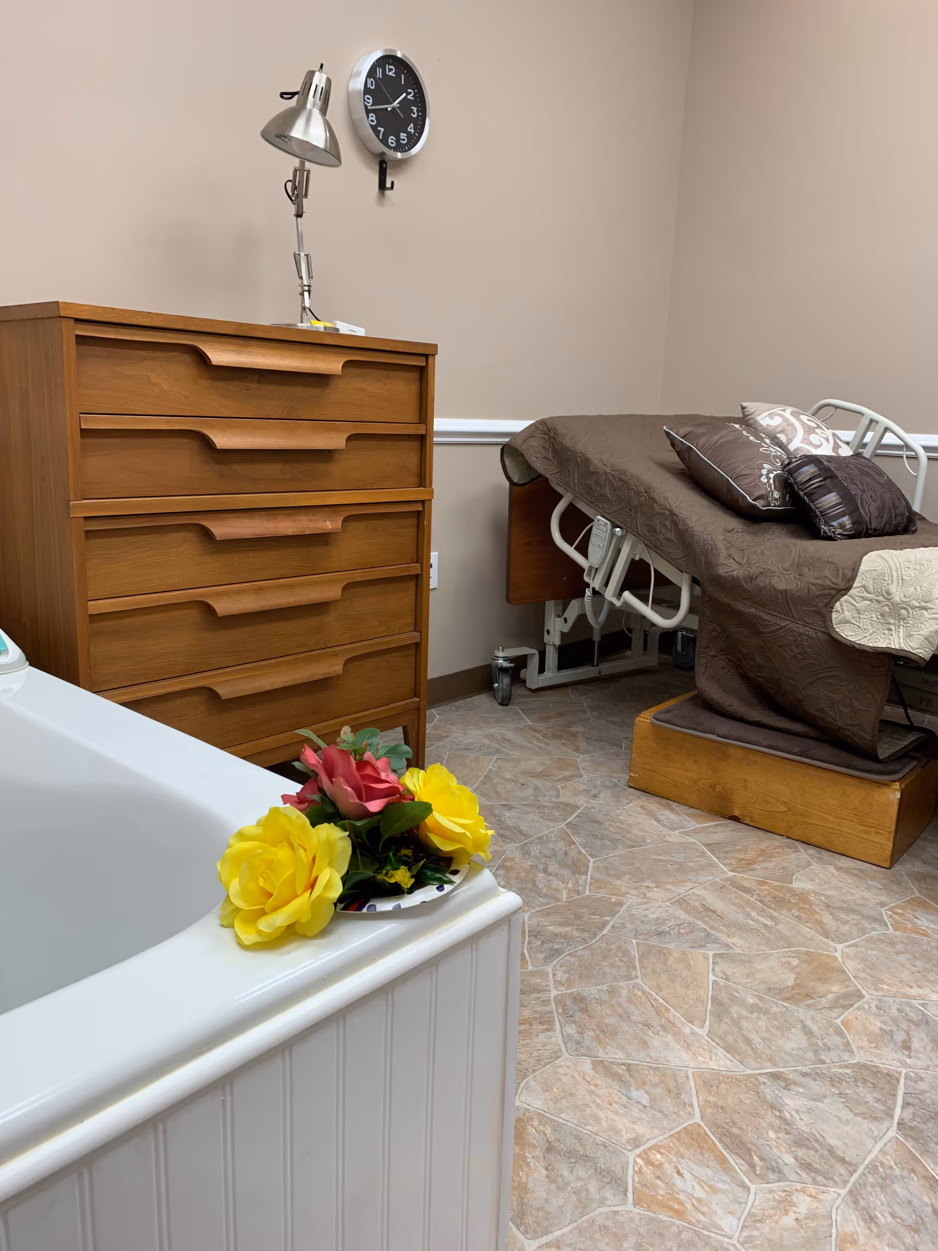 A hospital-style bed beside a wooden dresser in a beige-tiled care room with a bathtub.