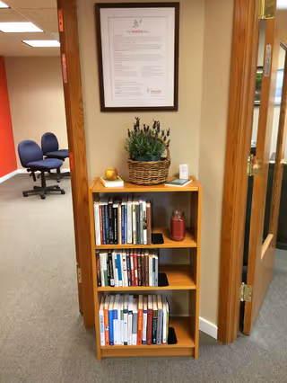 Wood bookshelf with books, plant, and framed notice in a hallway near open doors.