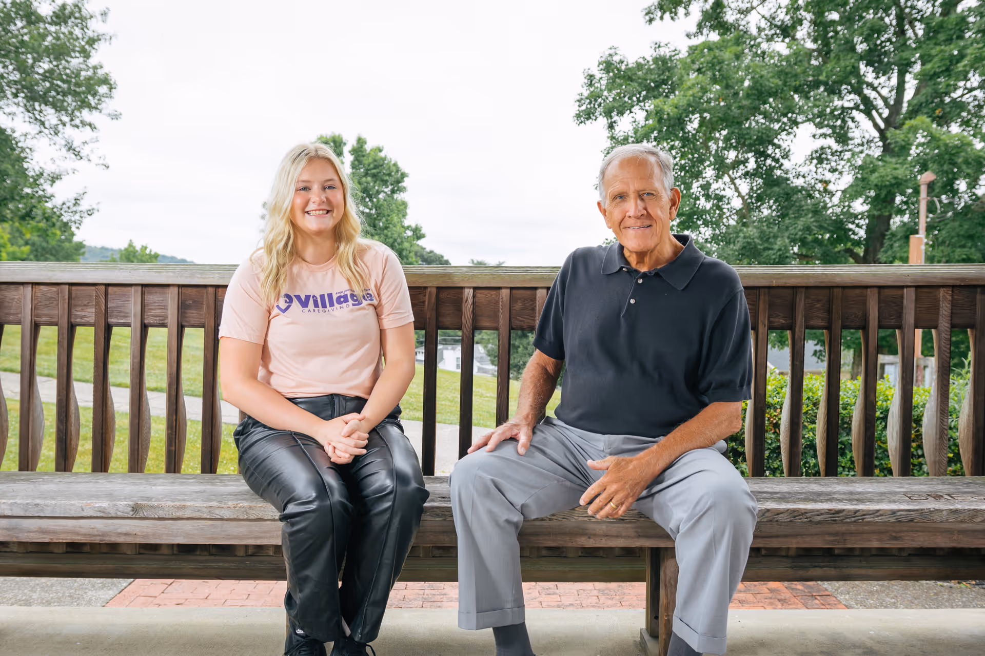 Two people sit on a wooden bench on an outdoor deck with trees in the background.