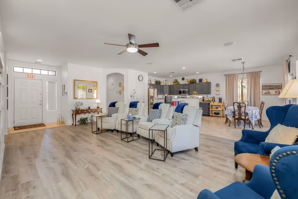 Spacious senior living lounge with white chairs, blue accents, and a kitchen beyond.