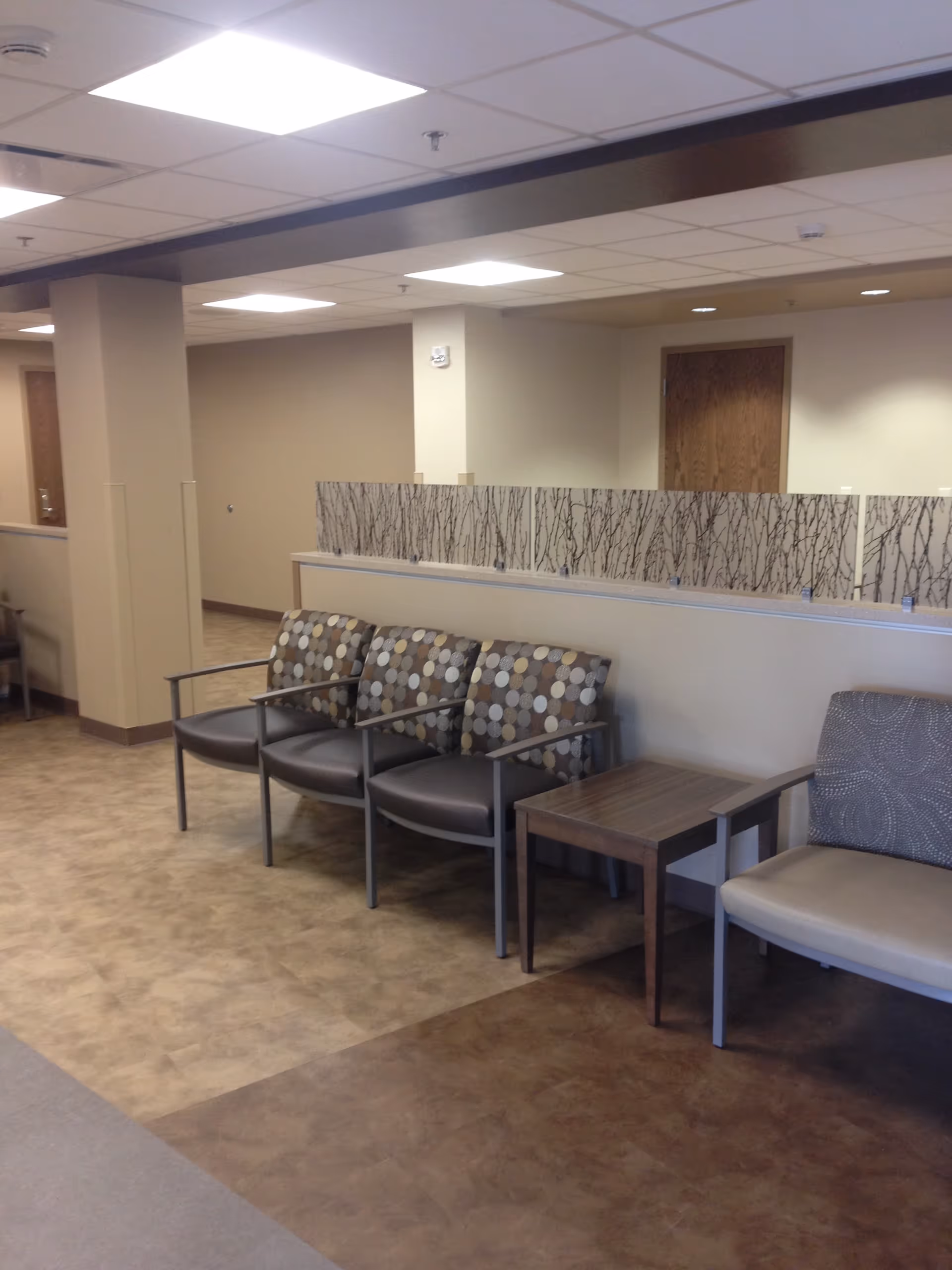 Waiting area with beige walls, tiled floor, and row of patterned chairs beside a partition.