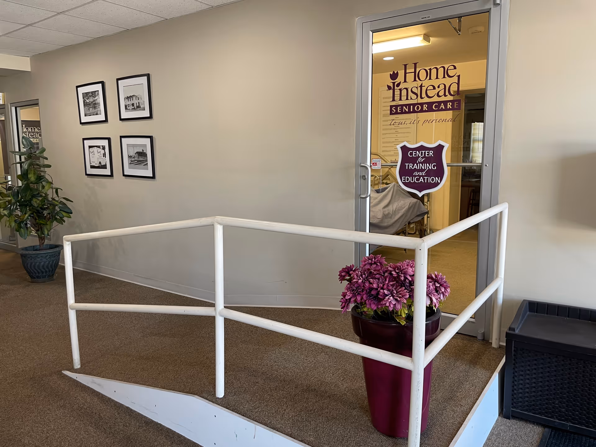 Interior hallway with ramp, framed photos, potted flowers, and a glass door labeled Home Instead.