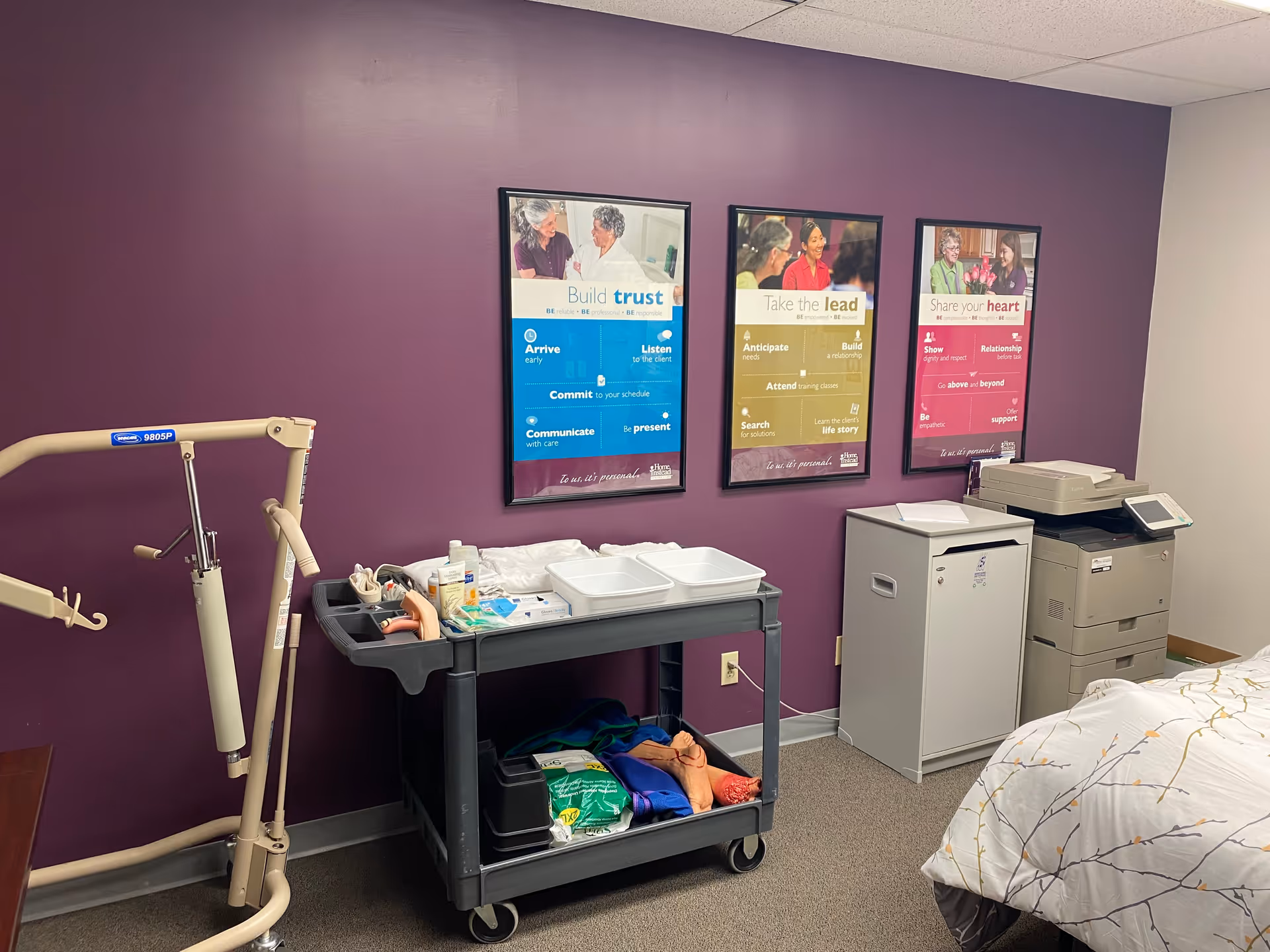 Purple-walled interior with a bed, medical cart, patient lift, and printer on a stand.