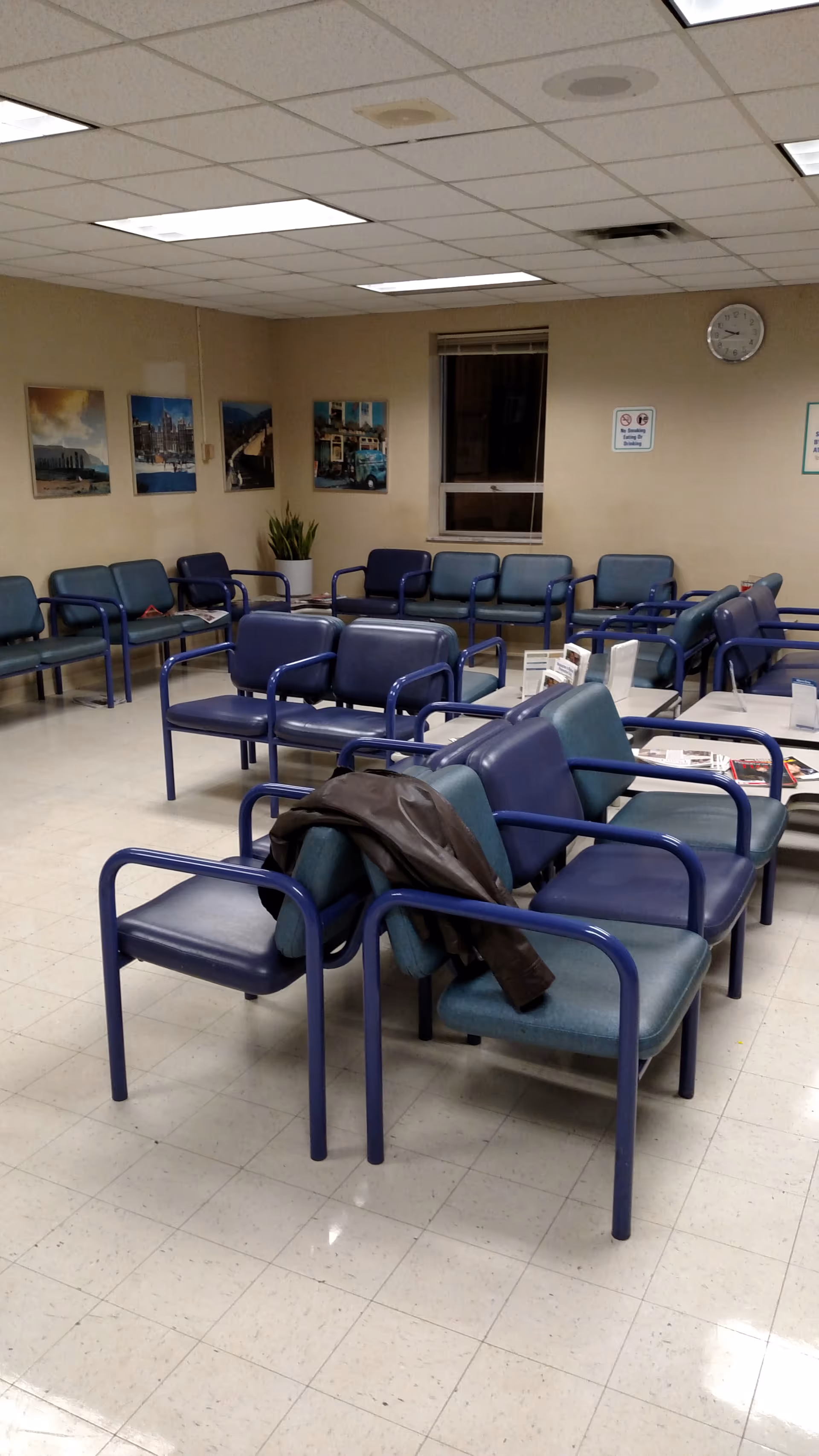 Interior waiting area with blue chairs, wall posters, a jacket on a chair, and fluorescent lighting.
