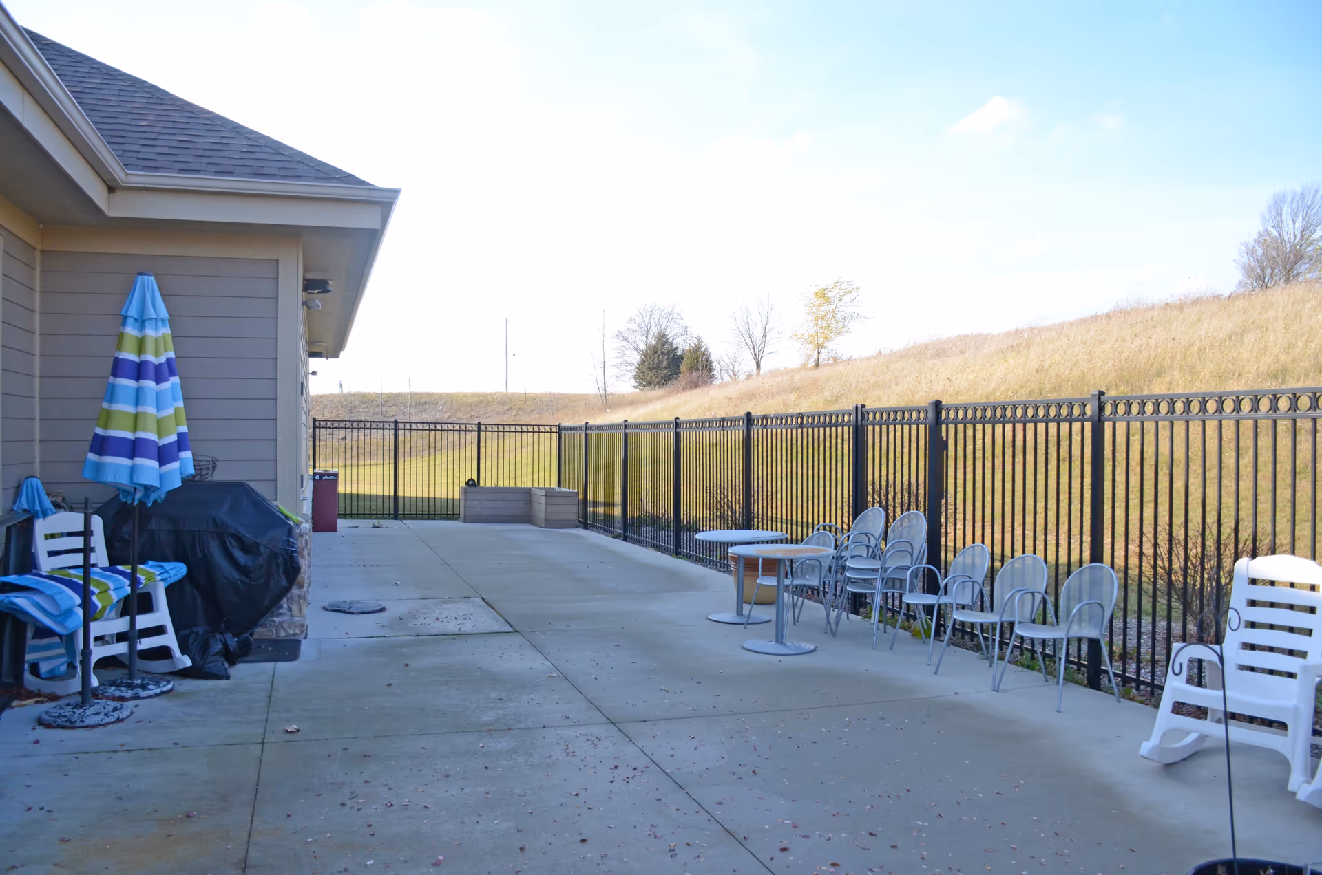 Outdoor concrete patio with chairs, tables, umbrellas, and a fence overlooking a grassy slope.