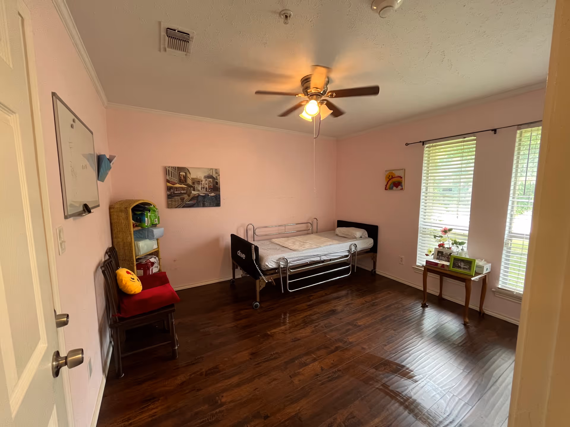 Pink-walled bedroom with hospital-style metal bed, ceiling fan, and wooden floor.