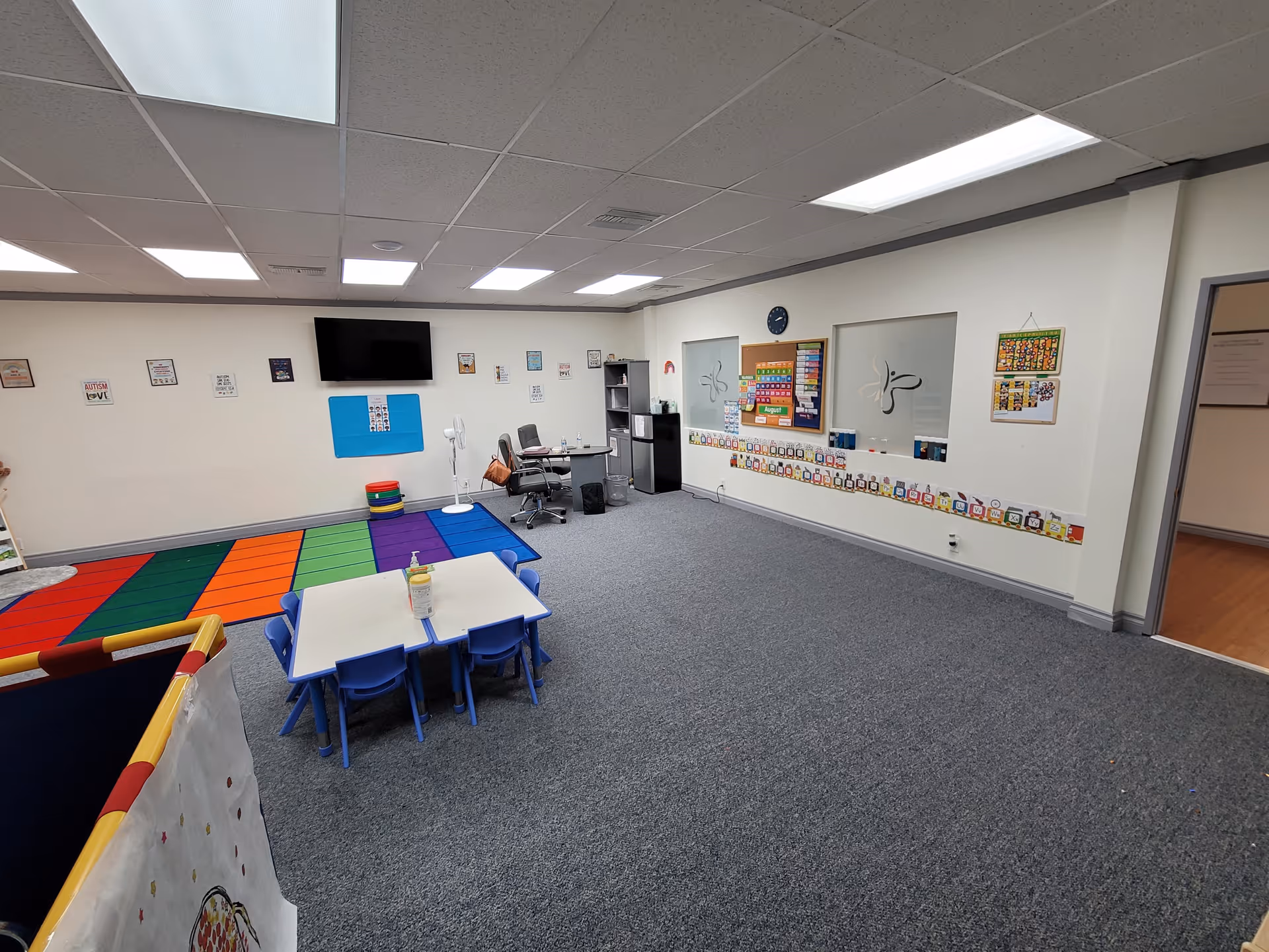 Bright activity room with colorful carpet squares, small tables, chairs, and wall displays.