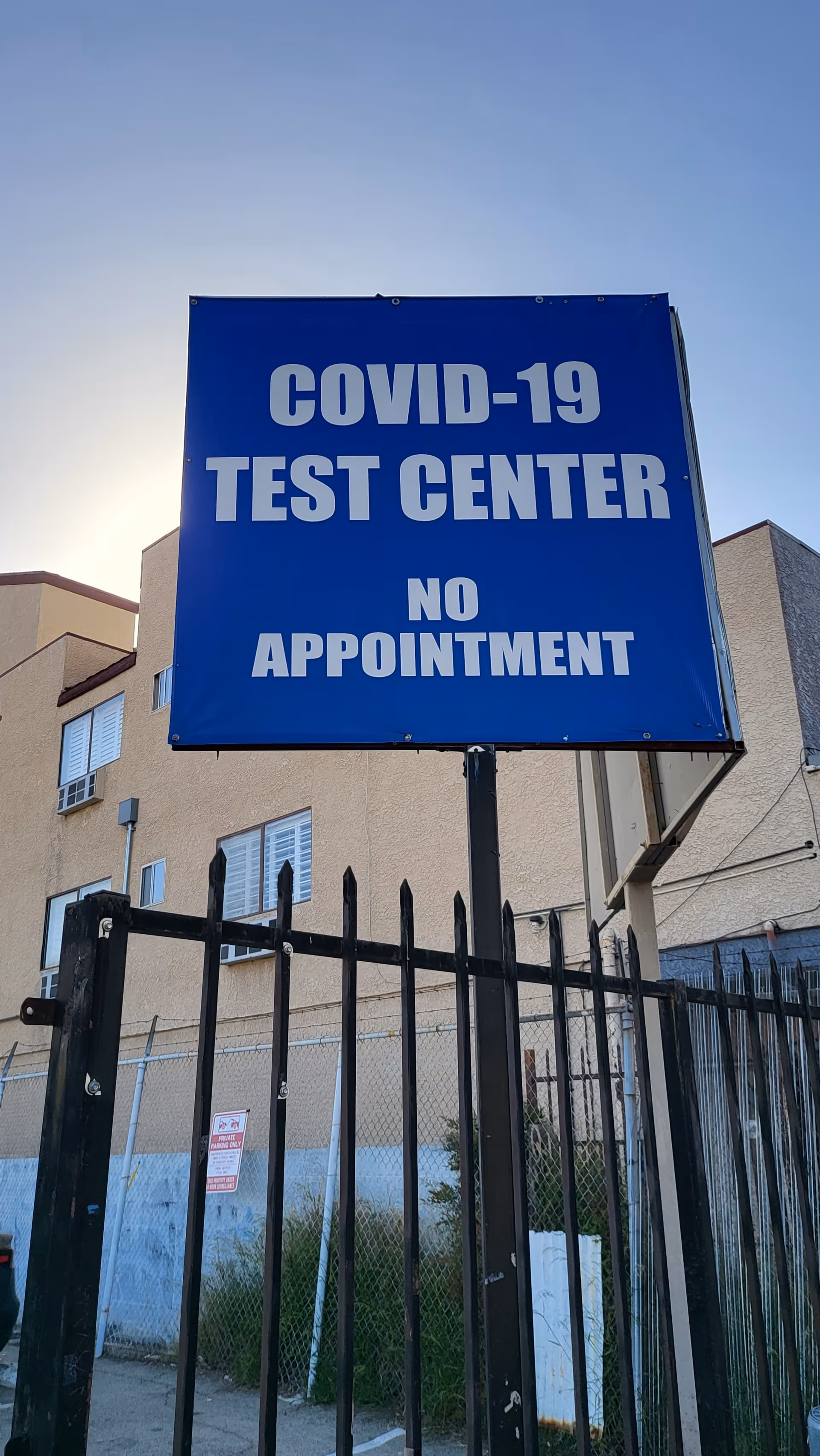 Blue COVID-19 test center sign behind a black fence and beige building.