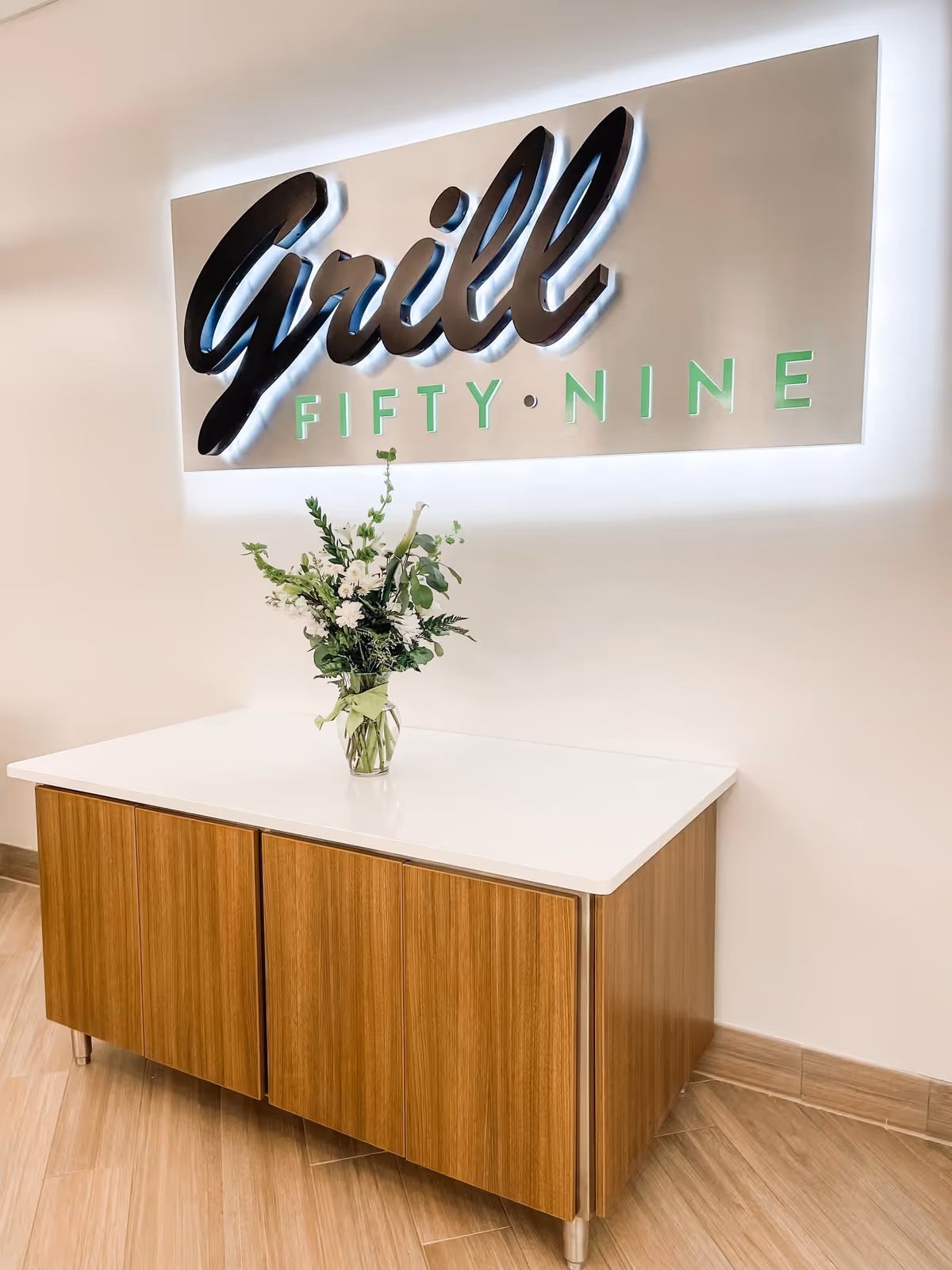 Reception area with a white countertop, wooden cabinet, and flower vase beneath a sign.