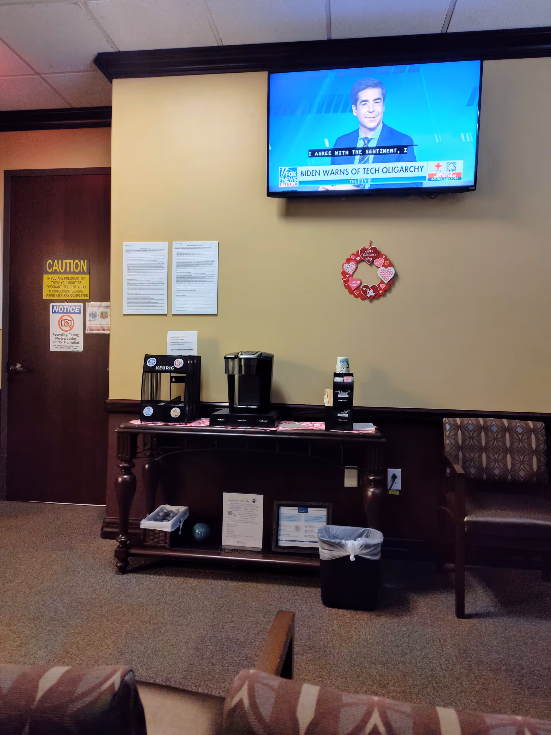 Indoor hospital lobby with coffee station, wall-mounted TV, and seating.