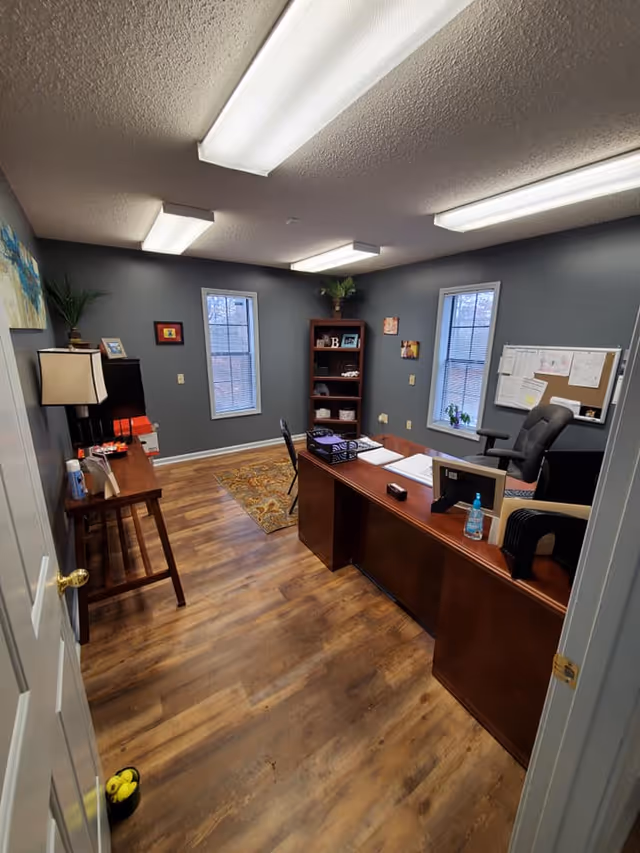 Office reception area with wooden desk, chairs, shelves, plants, and two windows in a senior facility.