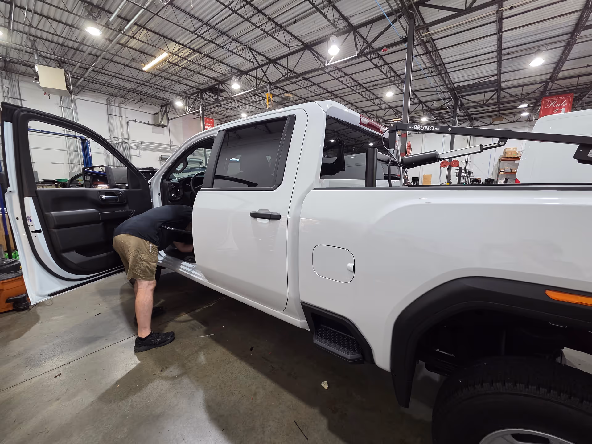 Man leans into the cab of a white pickup inside a spacious workshop.