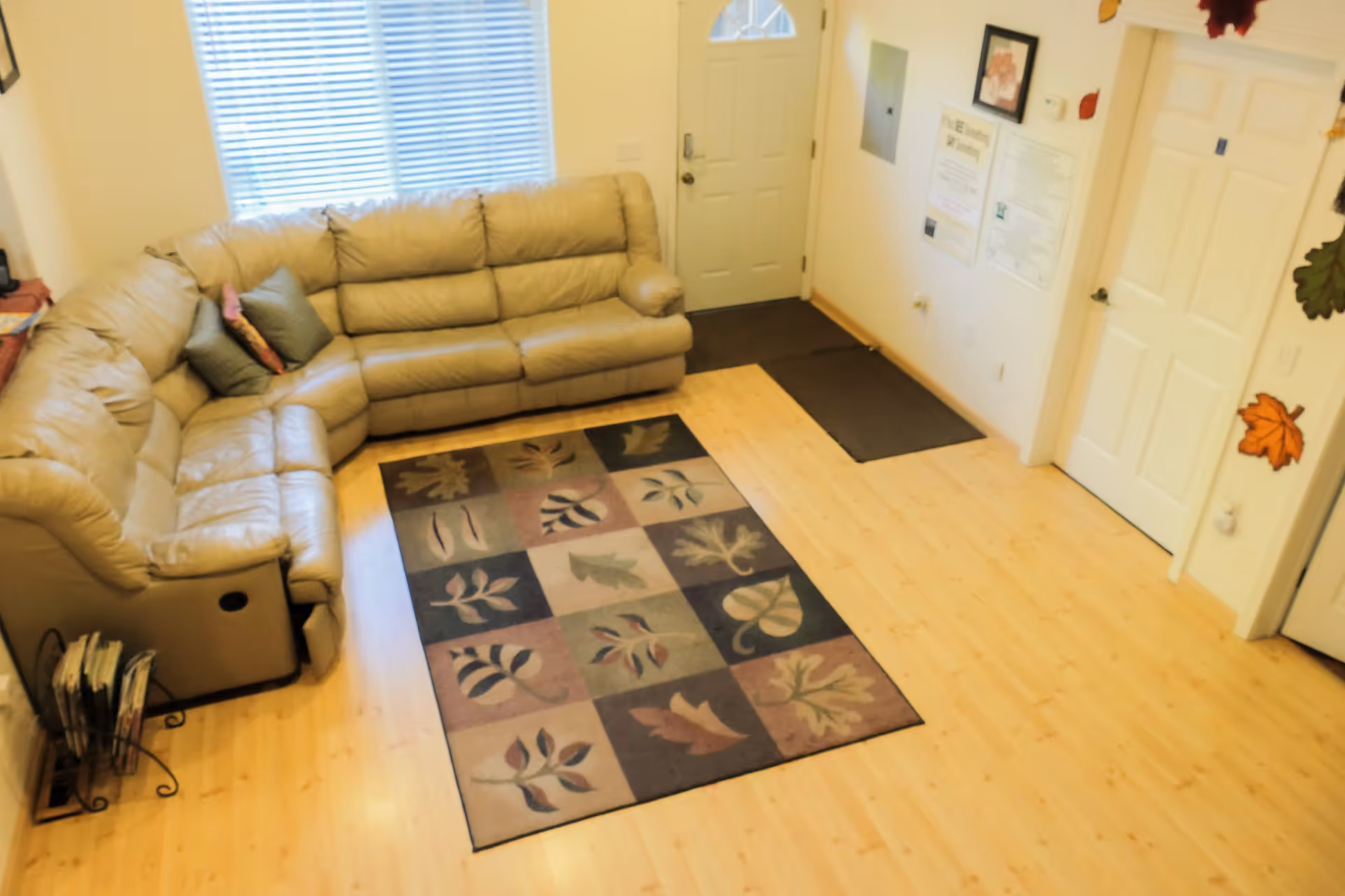 Bright living room with beige sectional sofa, window blinds, and a patterned leaf rug.