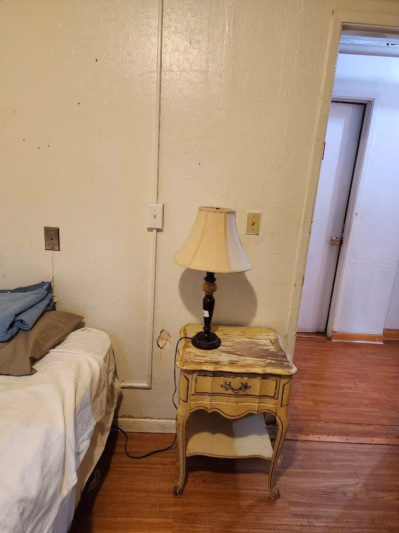 A small bedroom with a bed, worn nightstand, and a lamp beside the wall.