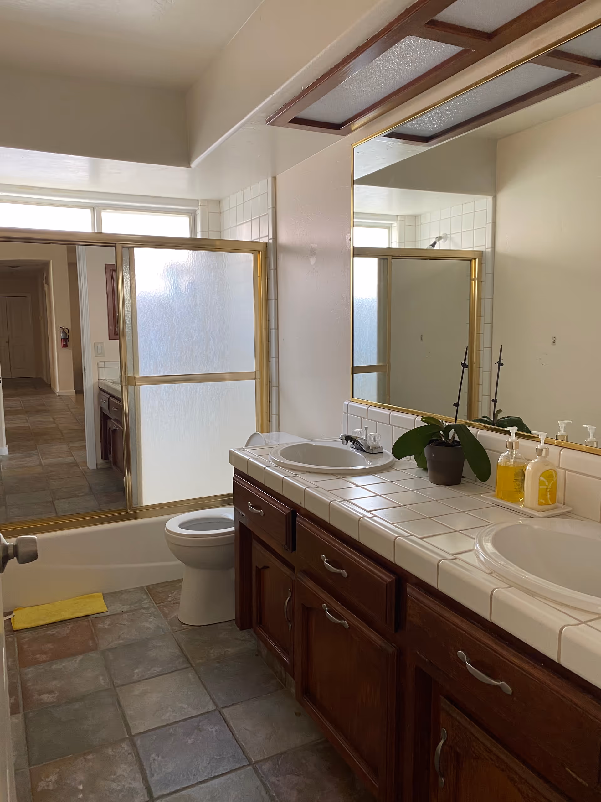 Bathroom with double sinks, tiled counter, toilet, and brass-framed frosted shower in a senior living facility.