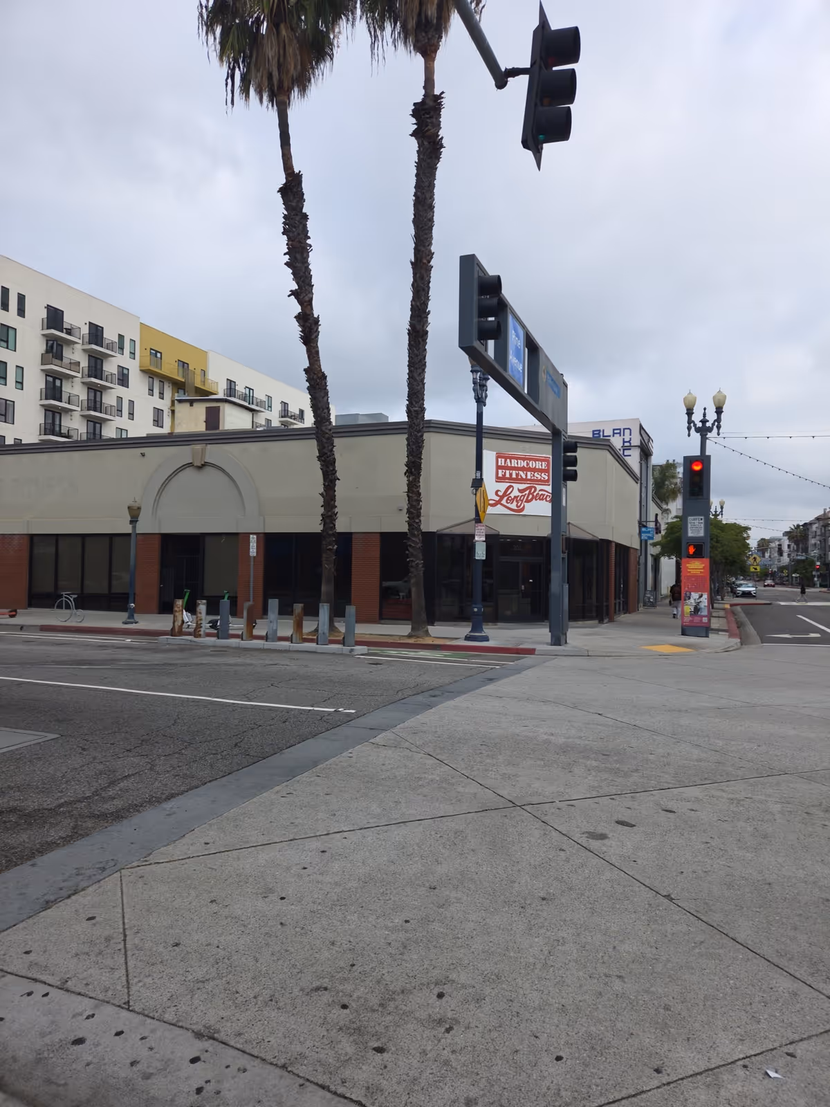 A street corner featuring a storefront, palm trees, traffic signals, and a wide sidewalk.