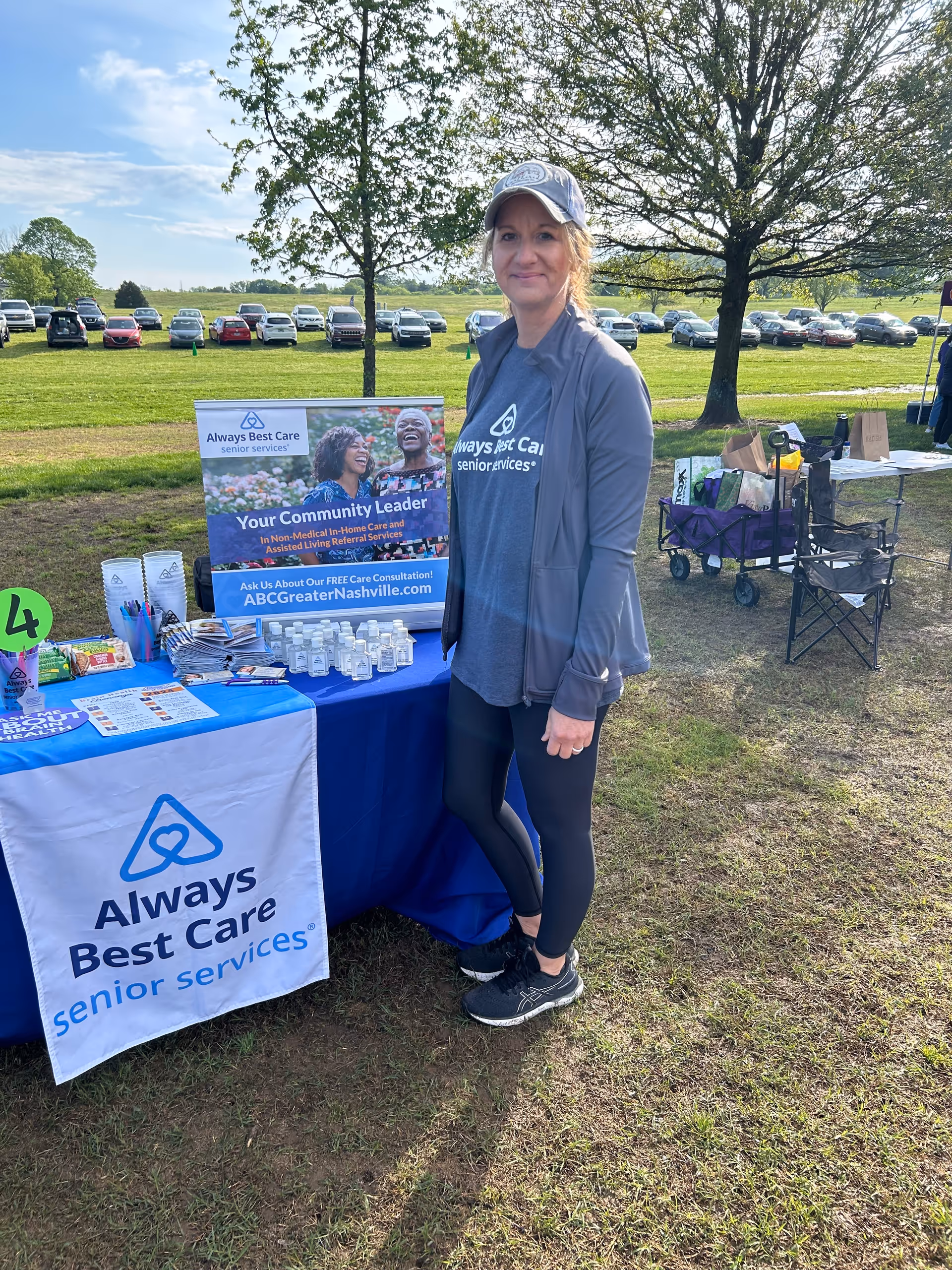 A woman stands beside an Always Best Care booth at an outdoor event.
