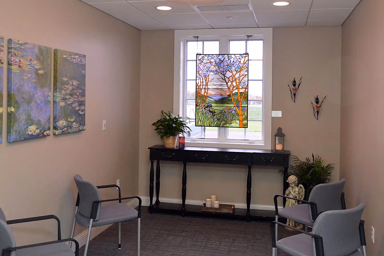 Small reception area with gray chairs, a dark console table, plants, and a stained glass window.