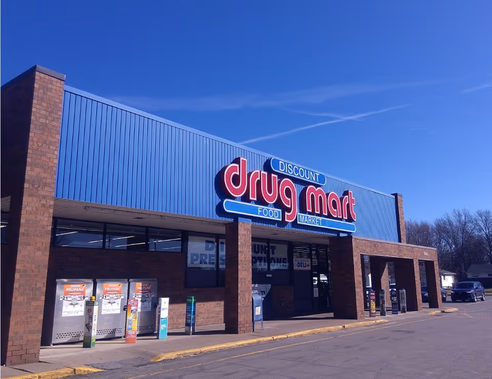 Brick storefront with bright blue awning and large red drug mart sign under a clear sky.
