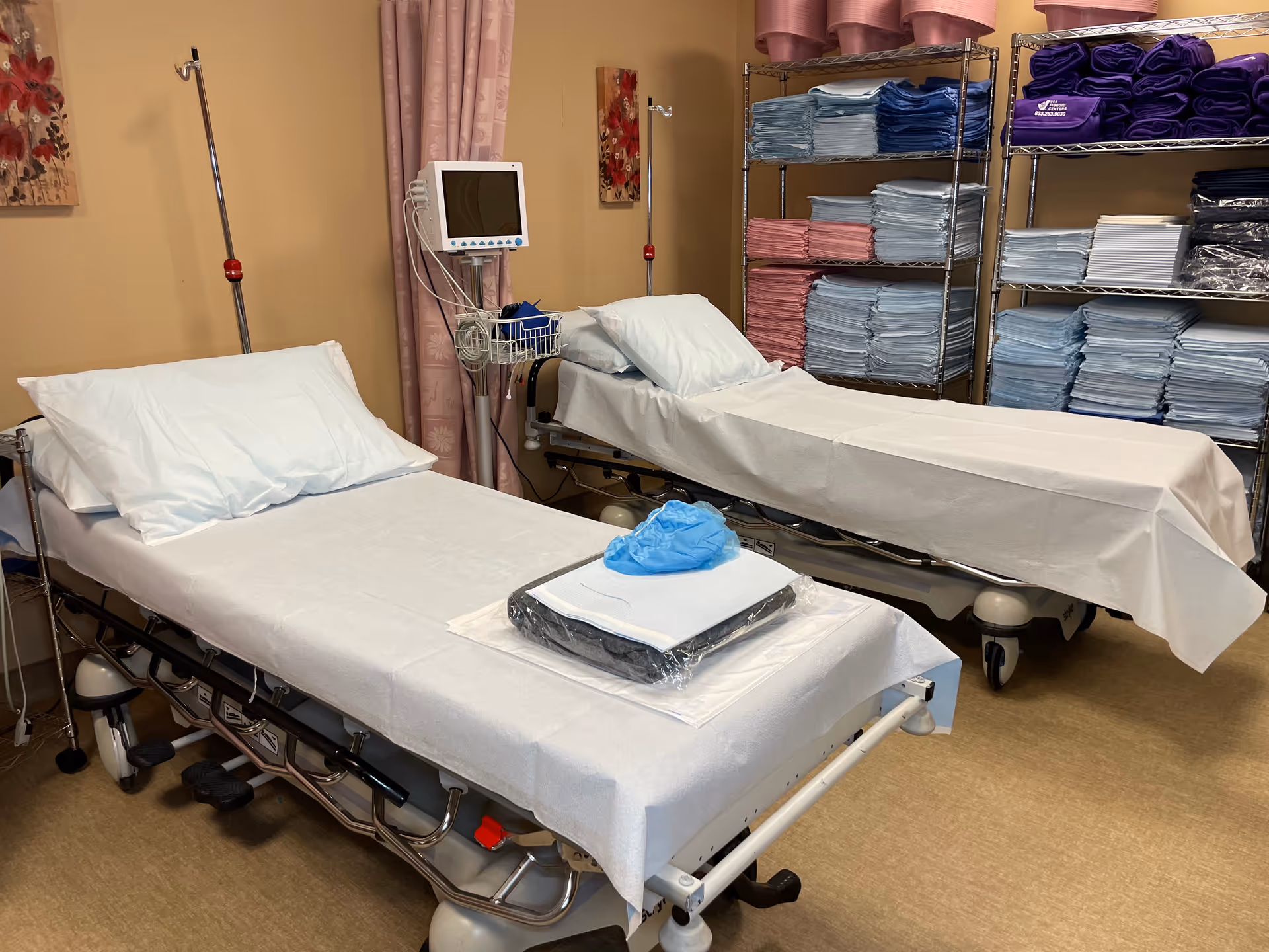 Two hospital beds with white sheets in a clinical procedure room, shelves of towels nearby.