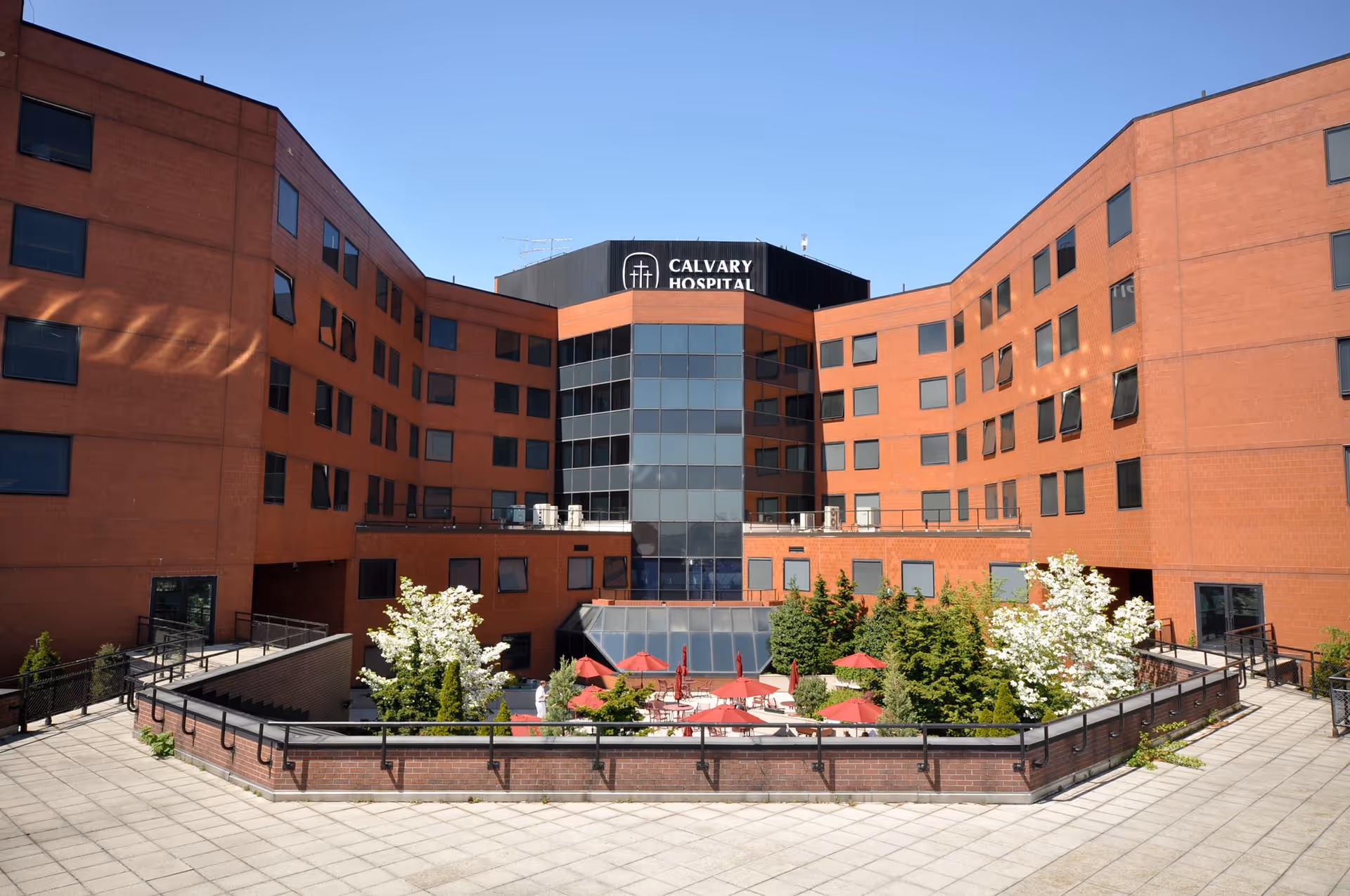 Exterior of Calvary Hospital: red-brick building, glass atrium, and courtyard with red umbrellas.