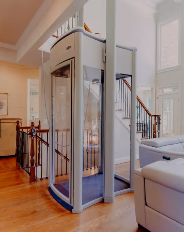 A compact residential elevator installed inside a bright living area with stairs and seating.