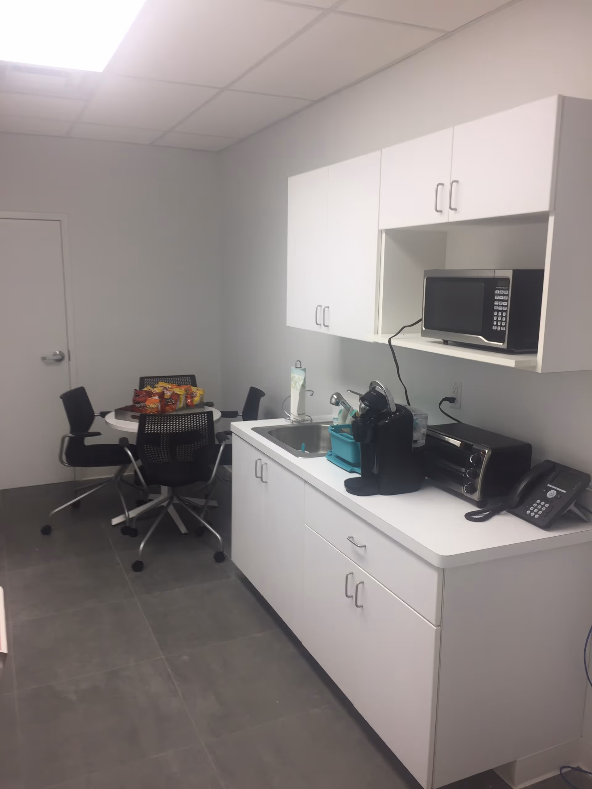 White kitchen with cabinets, sink, microwave, coffee machine, snacks, and a small round table with chairs.